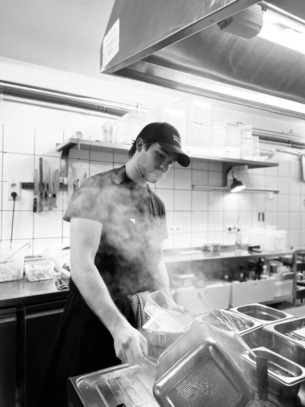 Un jeune homme en t-shirt noir et casquette travaille dans une cuisine de restaurant, soulevant un couvercle sur une casserole, avec de la vapeur qui s'en échappe, en noir et blanc.