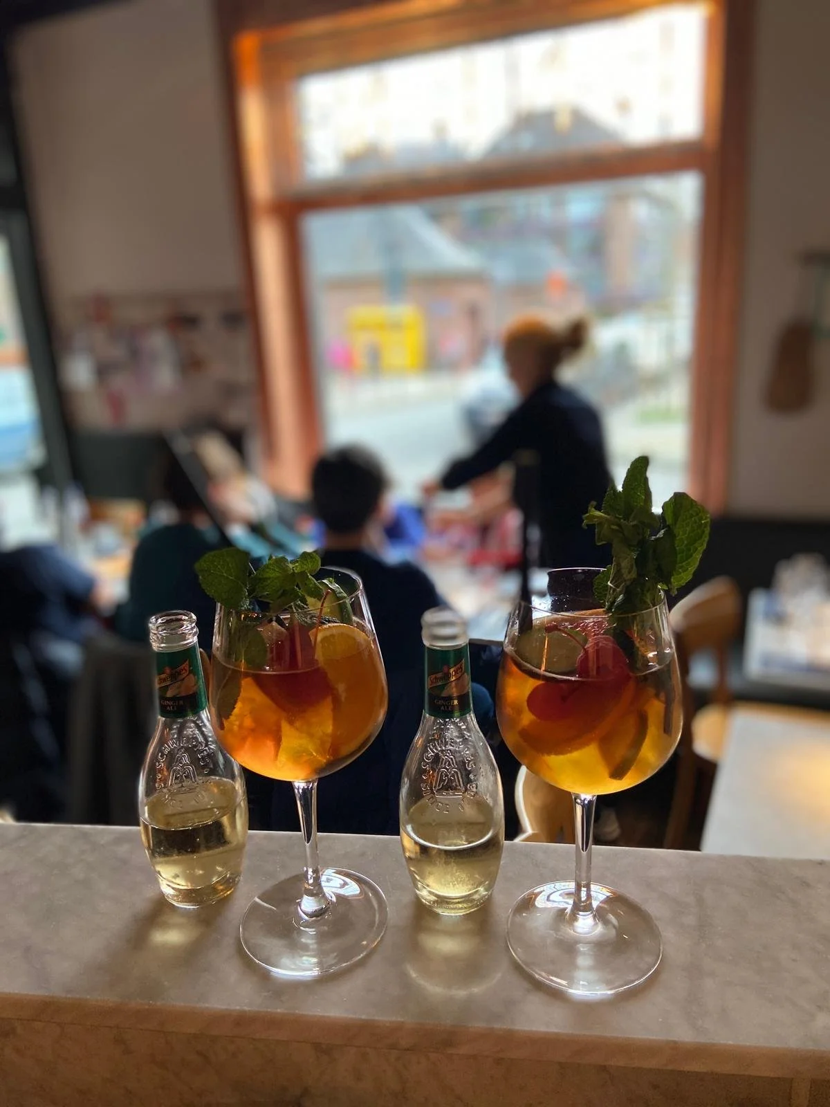 Deux verres de cocktail avec des feuilles de menthe, des cerises et des tranches de citron, accompagnés de deux petites bouteilles de soda au gingembre sur un comptoir de bar, dans un intérieur de restaurant ou café avec des personnes assises et une grande fenêtre en arrière-plan.