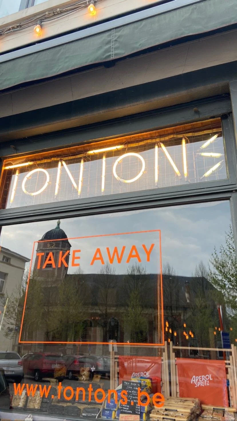 Vitrine d'un restaurant avec un néon allumé affichant le nom 'TON TON', un panneau indiquant 'TAKE AWAY', et l'adresse web 'www.tontons.be'. Reflection des bâtiments et arbres à l'extérieur.