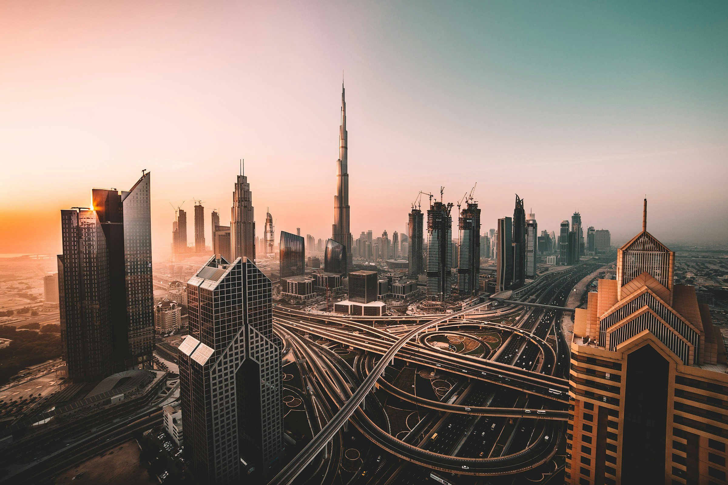 Skyline of Dubai with the Burj Khalifa in the center, numerous skyscrapers under construction, and busy highways with looping on-ramps at sunset.