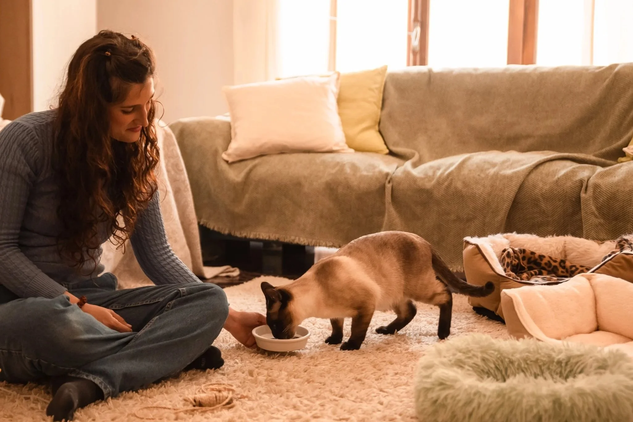 Una donna con capelli ricci lunghi si trova seduta sul tappeto in salotto accanto a un gatto siamese mentre mangia da una ciotola