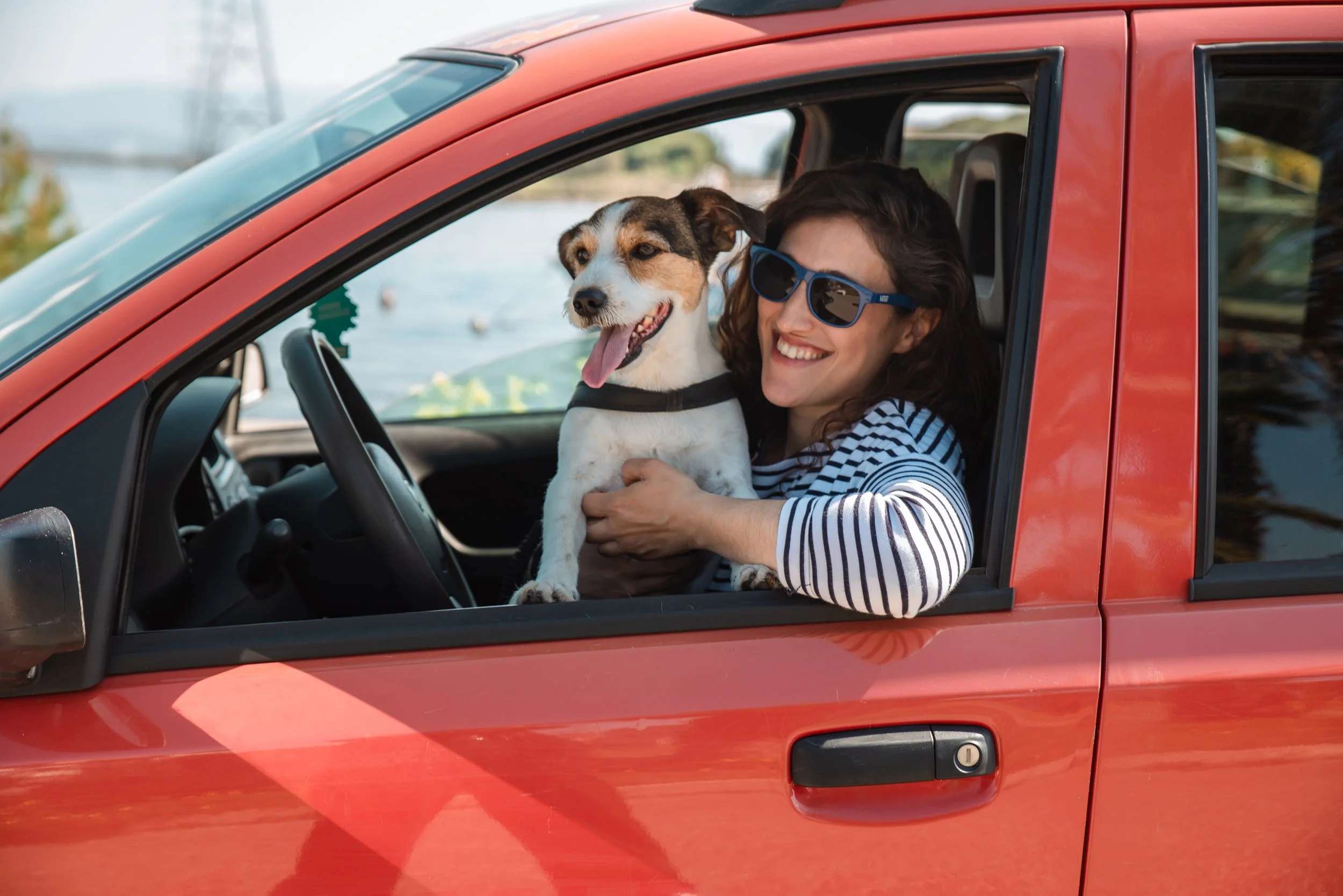 Una donna con occhiali da sole sorride e tiene un cane in braccio, seduta nel sedile di una macchina rossa vicino a un lago o riva.
