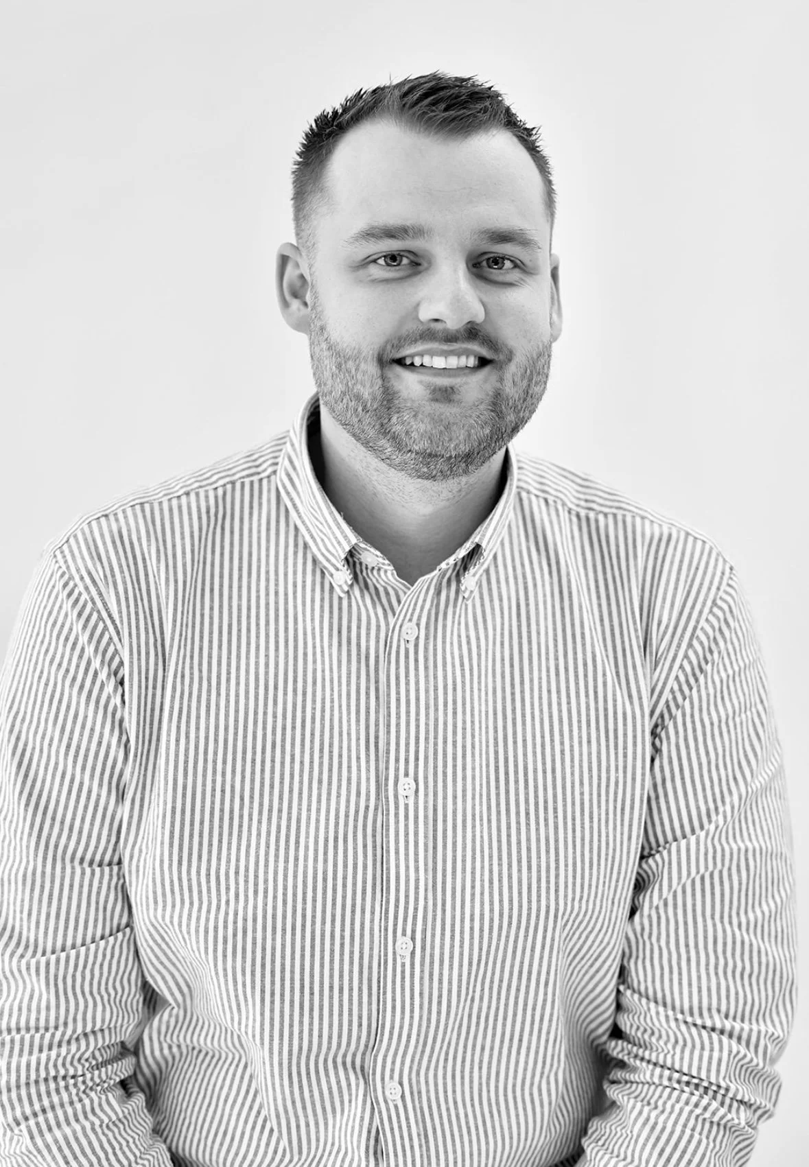 Black and white headshot of a smiling man with short hair and beard wearing a striped collared shirt.