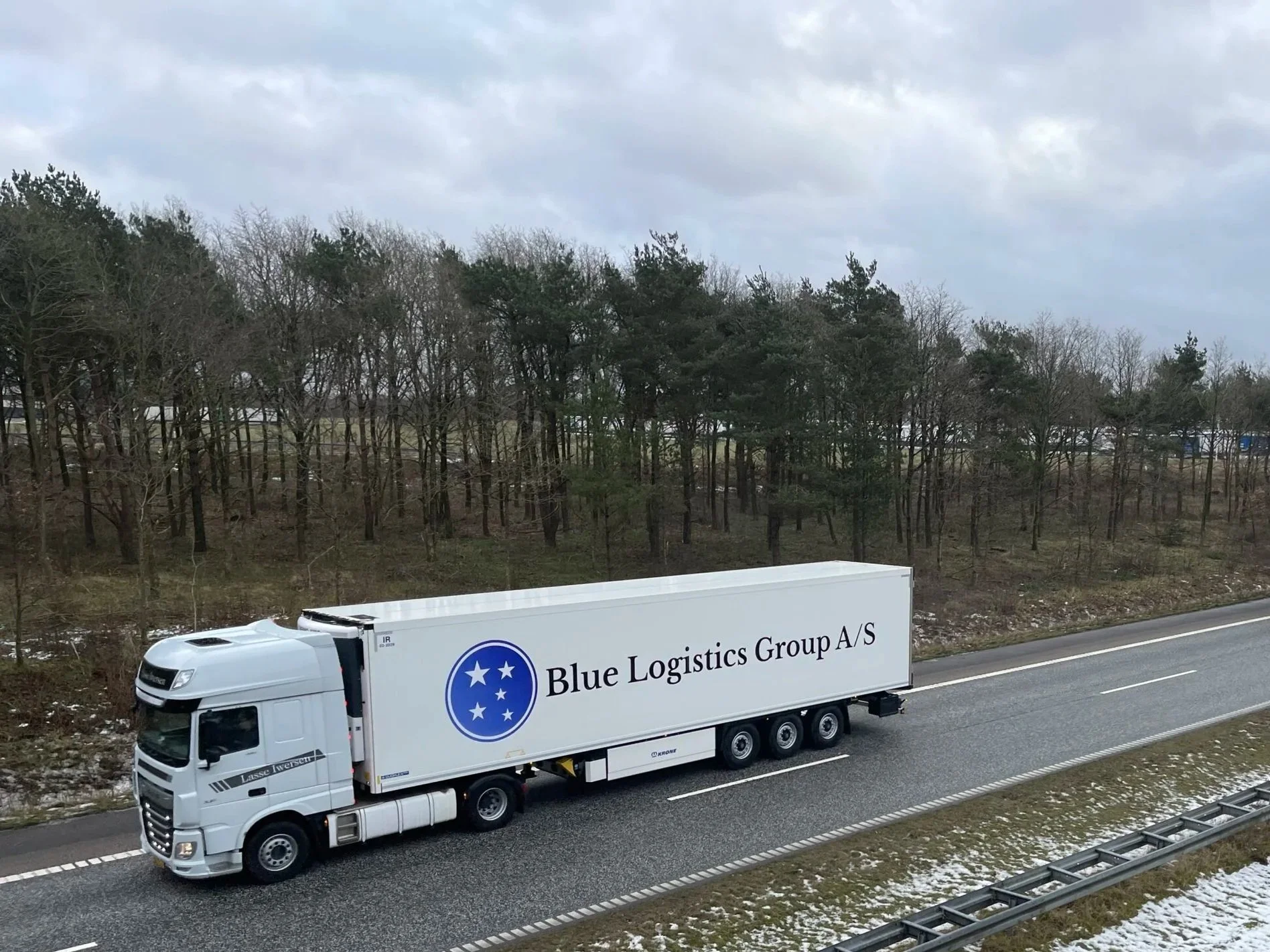 A white semi-truck with a trailer labeled 'Blue Logistics Group A/S' driving on a highway near a wooded area with overcast skies.