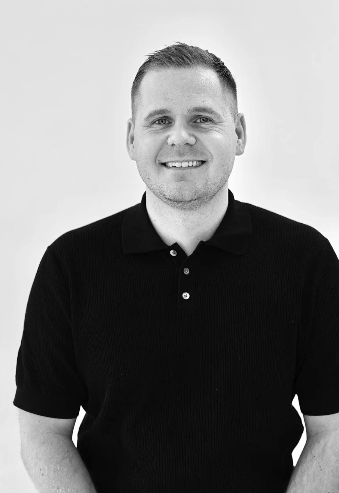 Black and white portrait of a smiling young man with short hair, wearing a black collared shirt with buttons against a plain light background.