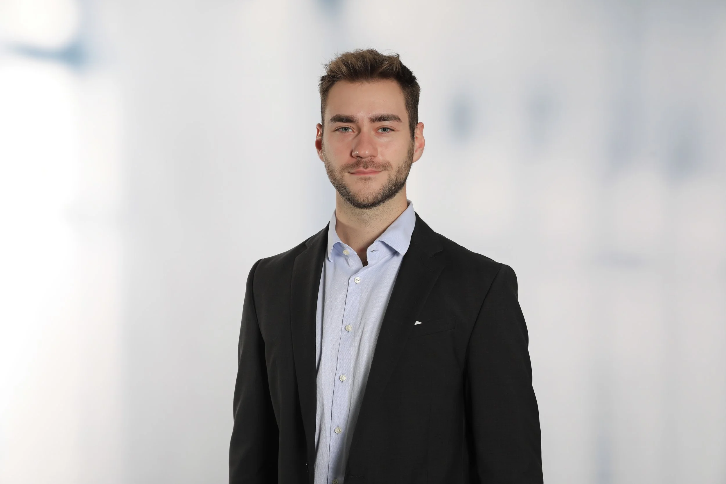 Junger Mann in dunklem Anzug und hellem Hemd vor neutralem Hintergrund.