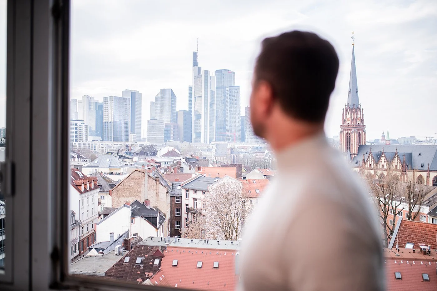 Ein Mann schaut durch ein offenes Fenster auf eine Skyline mit modernen Wolkenkratzern und einer historischen Kirche in Frankfurt, Deutschland.