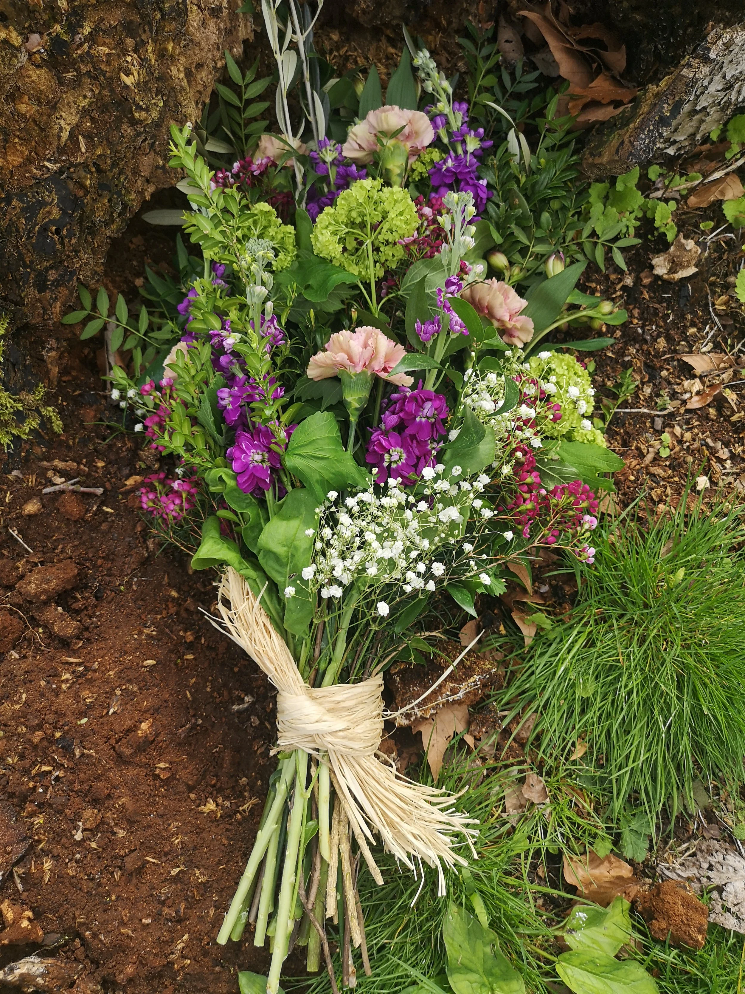 a purple bouquet rests on warm brown soil at the base of an oak tree. the bouquet is tied with flaxen thread.