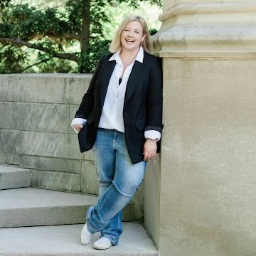A young woman with blonde hair smiling while leaning against a stone column outdoors, wearing a black blazer, white shirt, and jeans