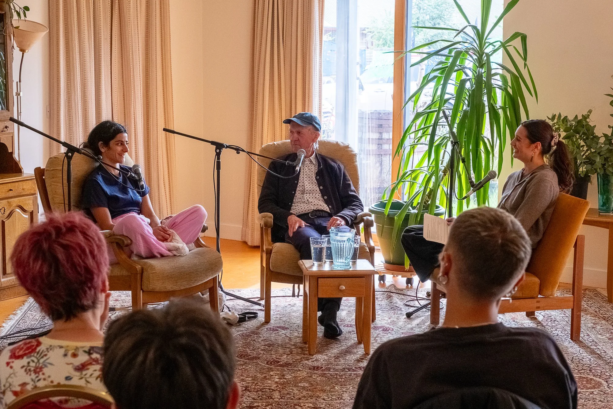 A panel discussion with two women and one man seated in a cozy room with large plants, sunlight, and an audience.