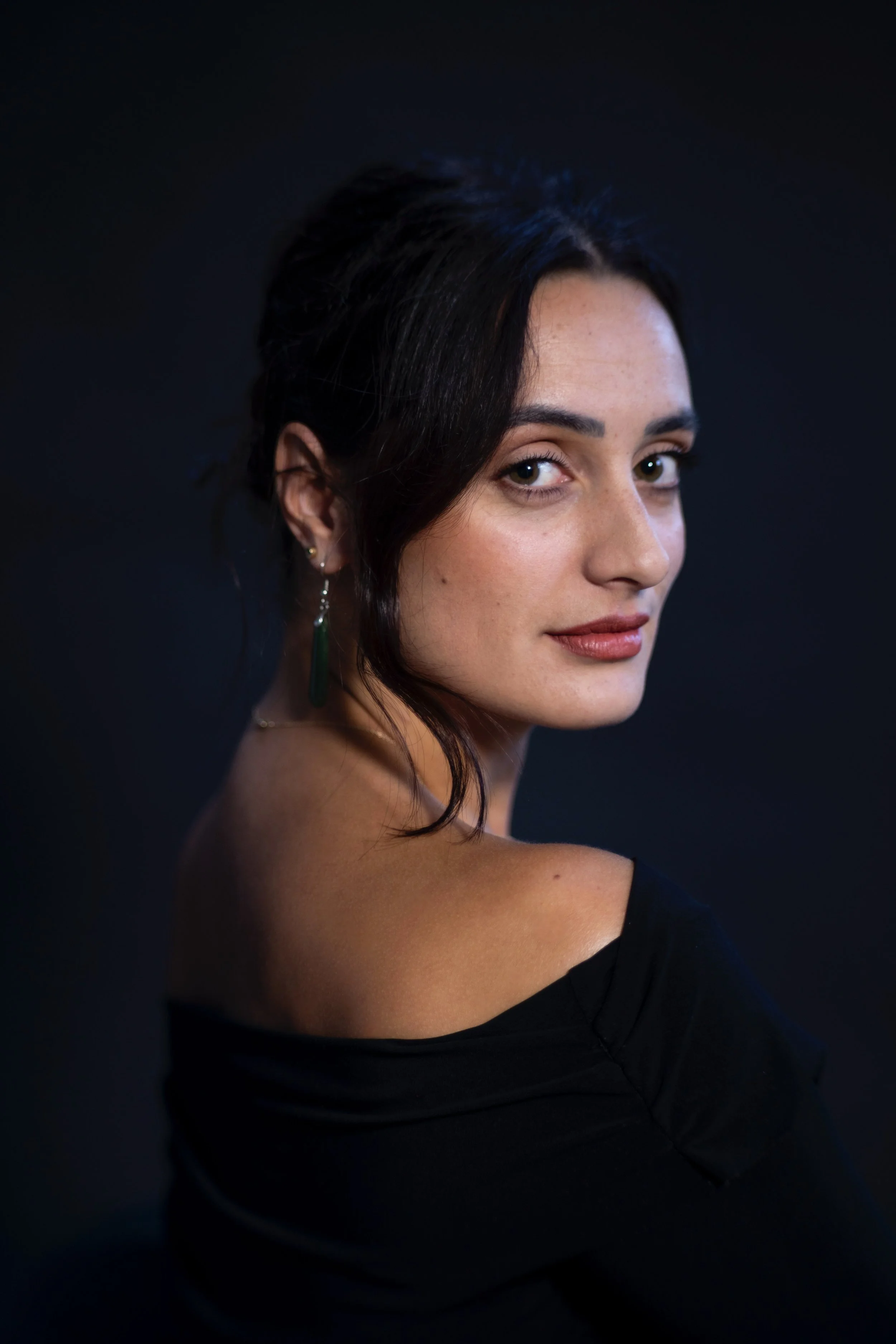 A woman with brown hair, wearing an olive green sweater, sitting and smiling near a microphone, holding a pen and some papers, in a well-lit room with large windows.