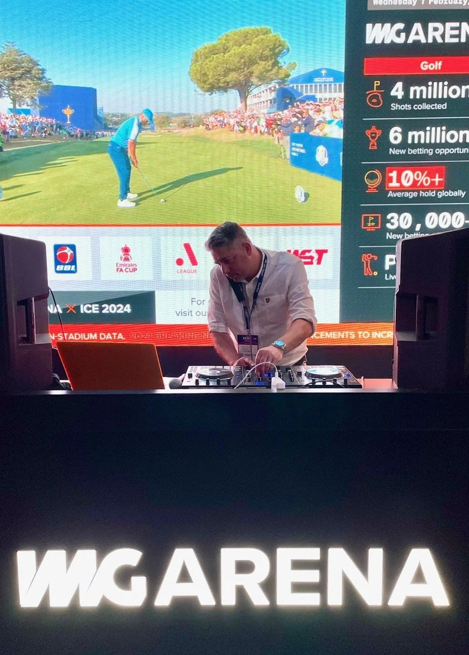 A man in a white shirt and lanyard, DJing at a booth with a large screen behind him showing a golf game and betting statistics at the MG Arena during an event.