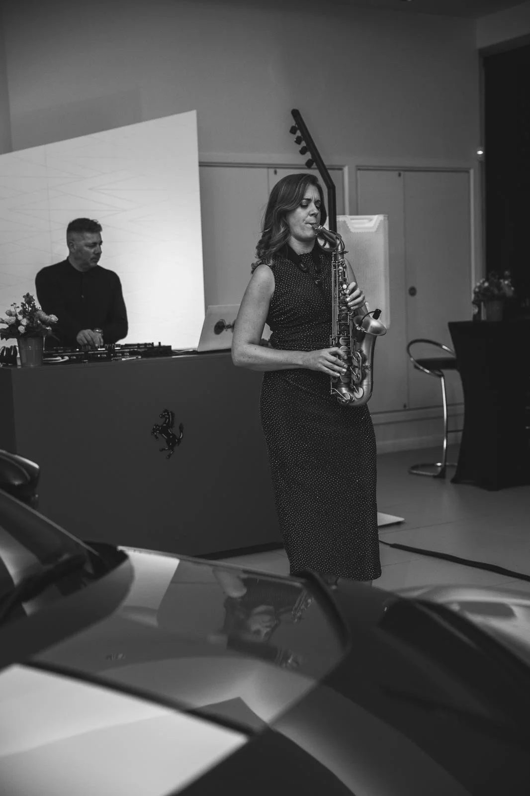 A woman playing a saxophone during a live performance with a DJ in the background at an indoor event.