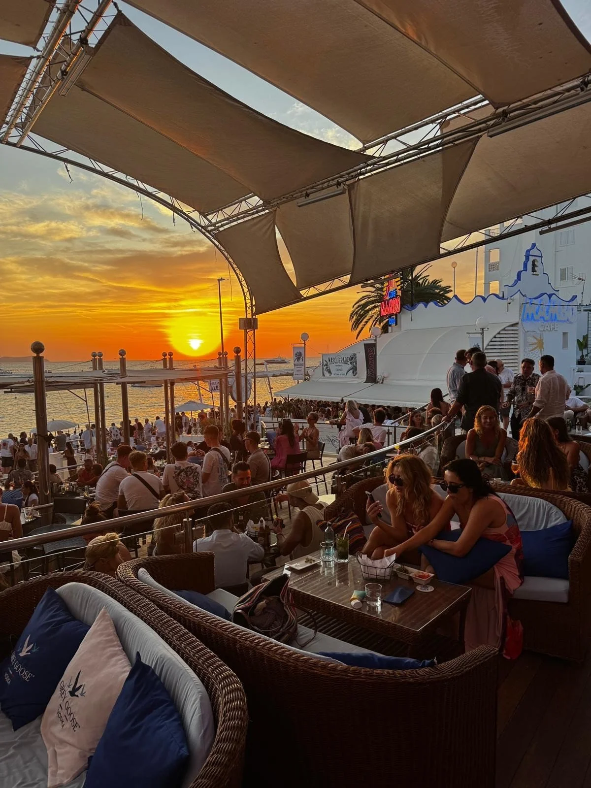 People dining and socializing at a beachside restaurant during sunset, with a view of the ocean and sunset in the background.