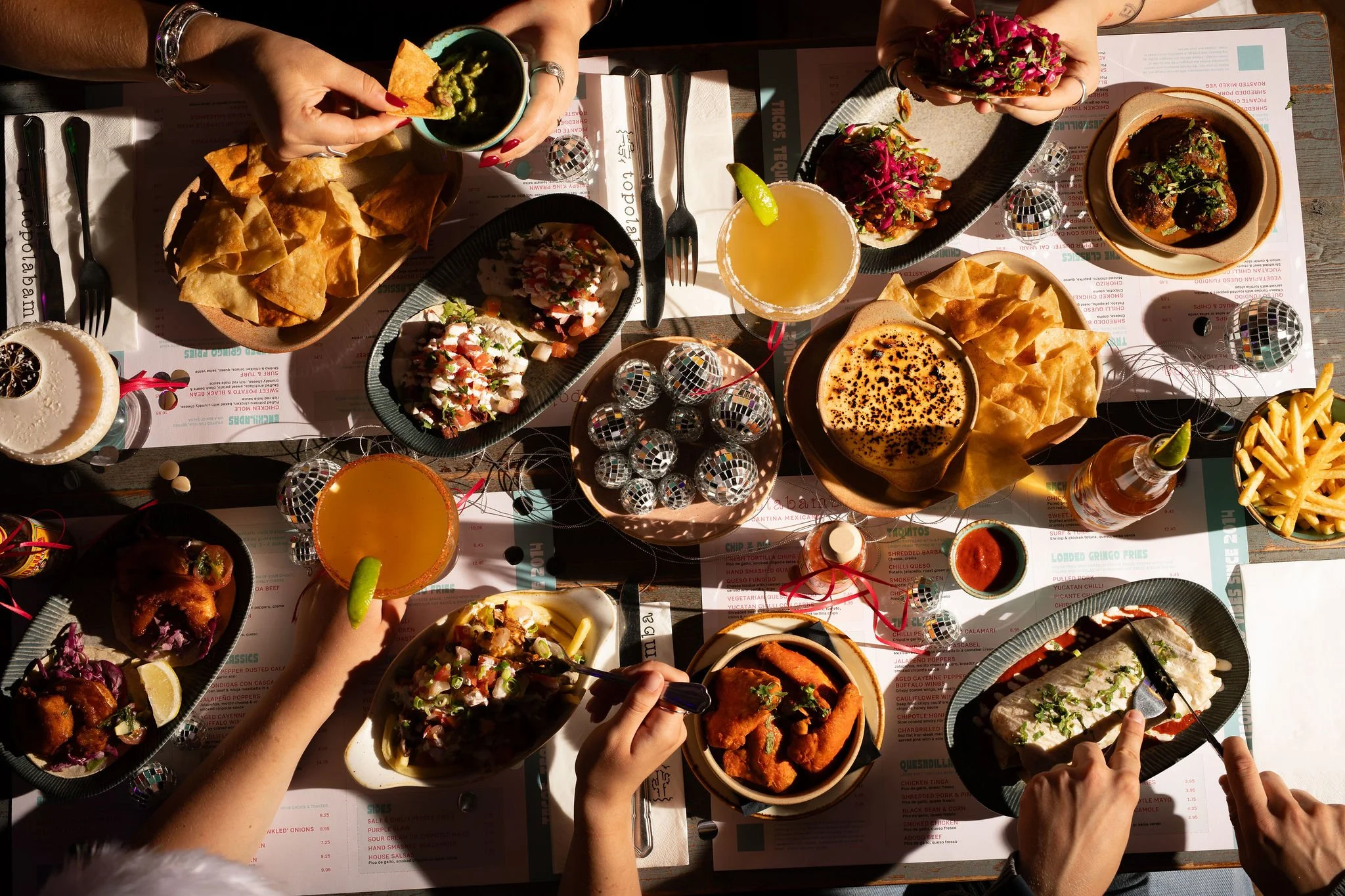 A table filled with Mexican food dishes including tacos, nachos, dips, and drinks, with hands reaching for food and utensils.
