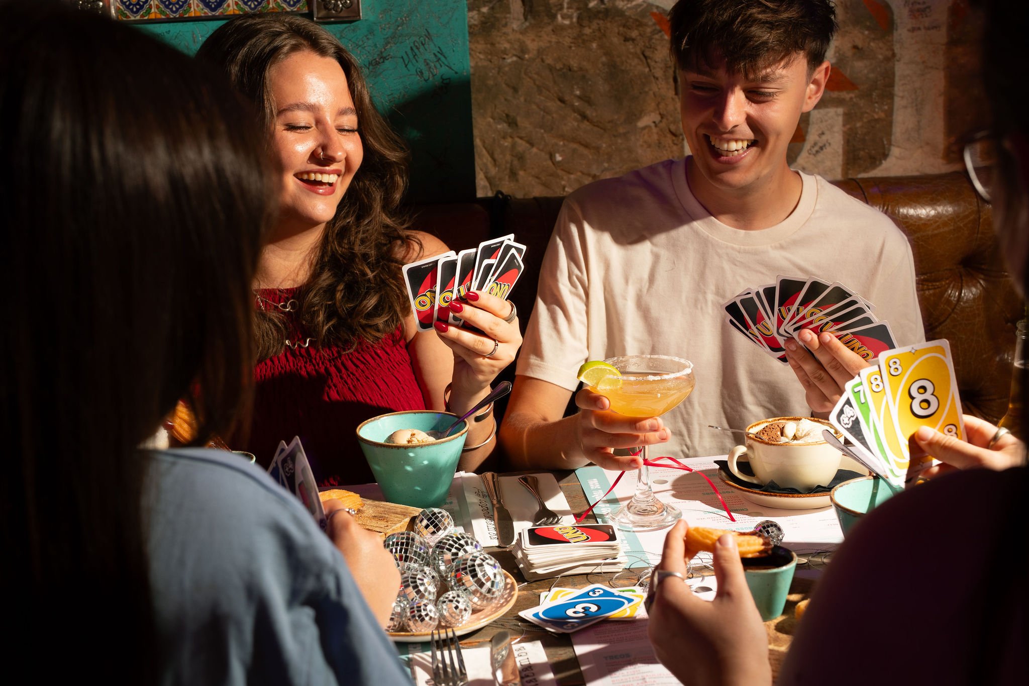 Group of friends playing Uno card game at a restaurant table with desserts and drinks.