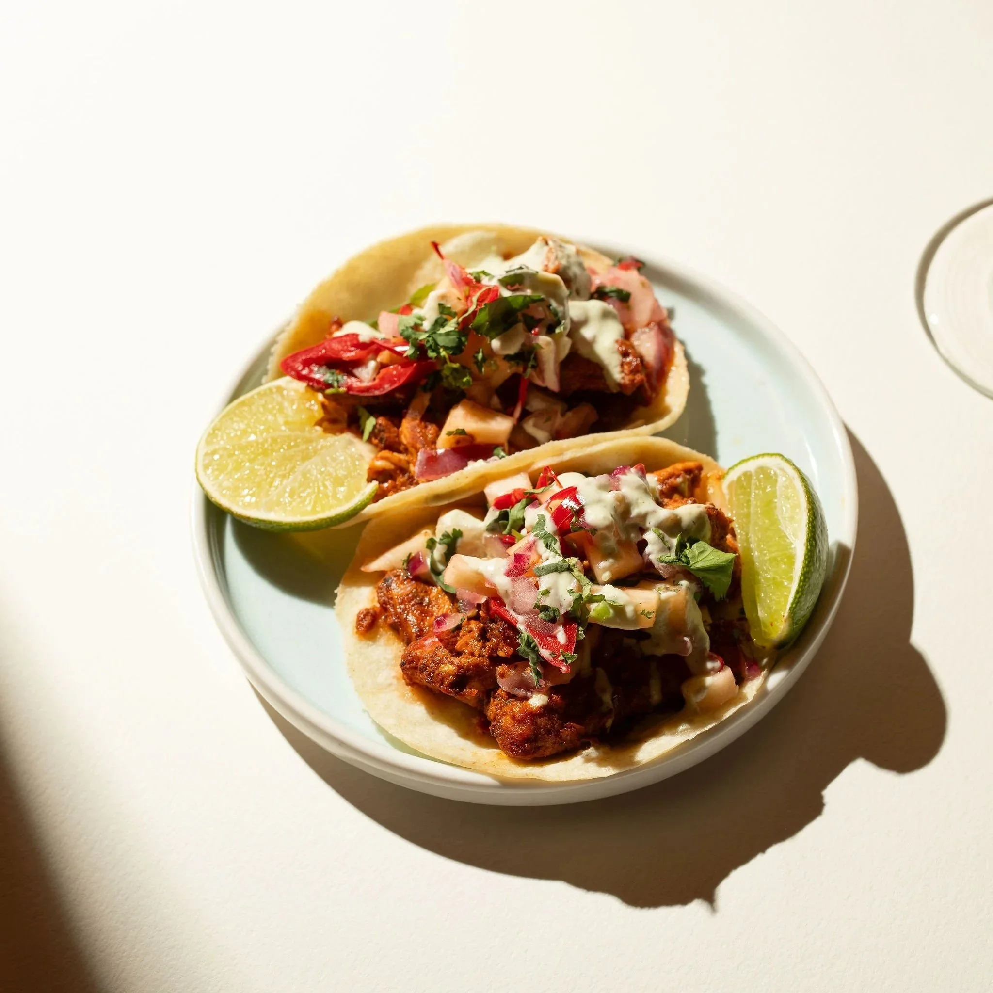 Close-up of three small tacos with red meat, onions, cilantro, and creamy sauce, garnished with lime wedges on a white plate.