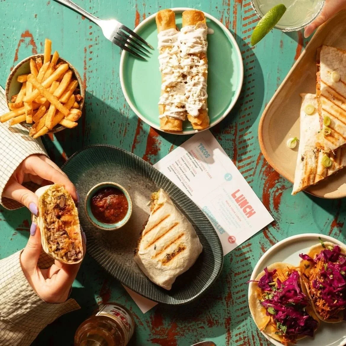 A variety of Mexican food including fries, burritos, quesadillas, tacos, and drinks on a turquoise wooden table.