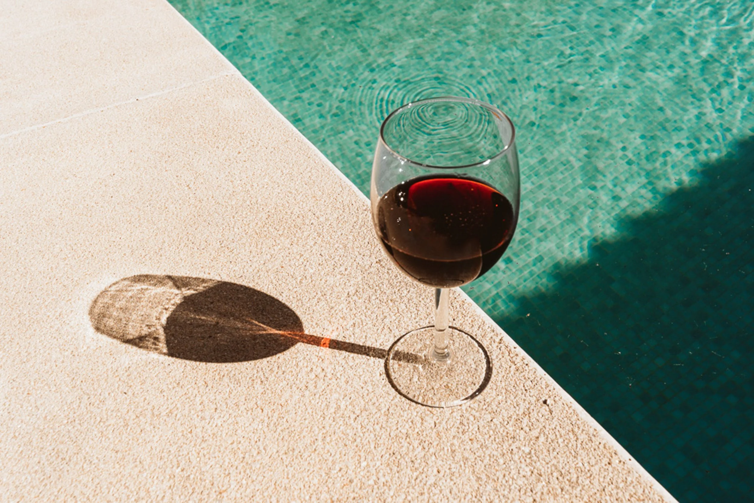 Ein Weinglas mit rotem Wein neben einem Pool, Schatten und Wasserreflexionen sichtbar.