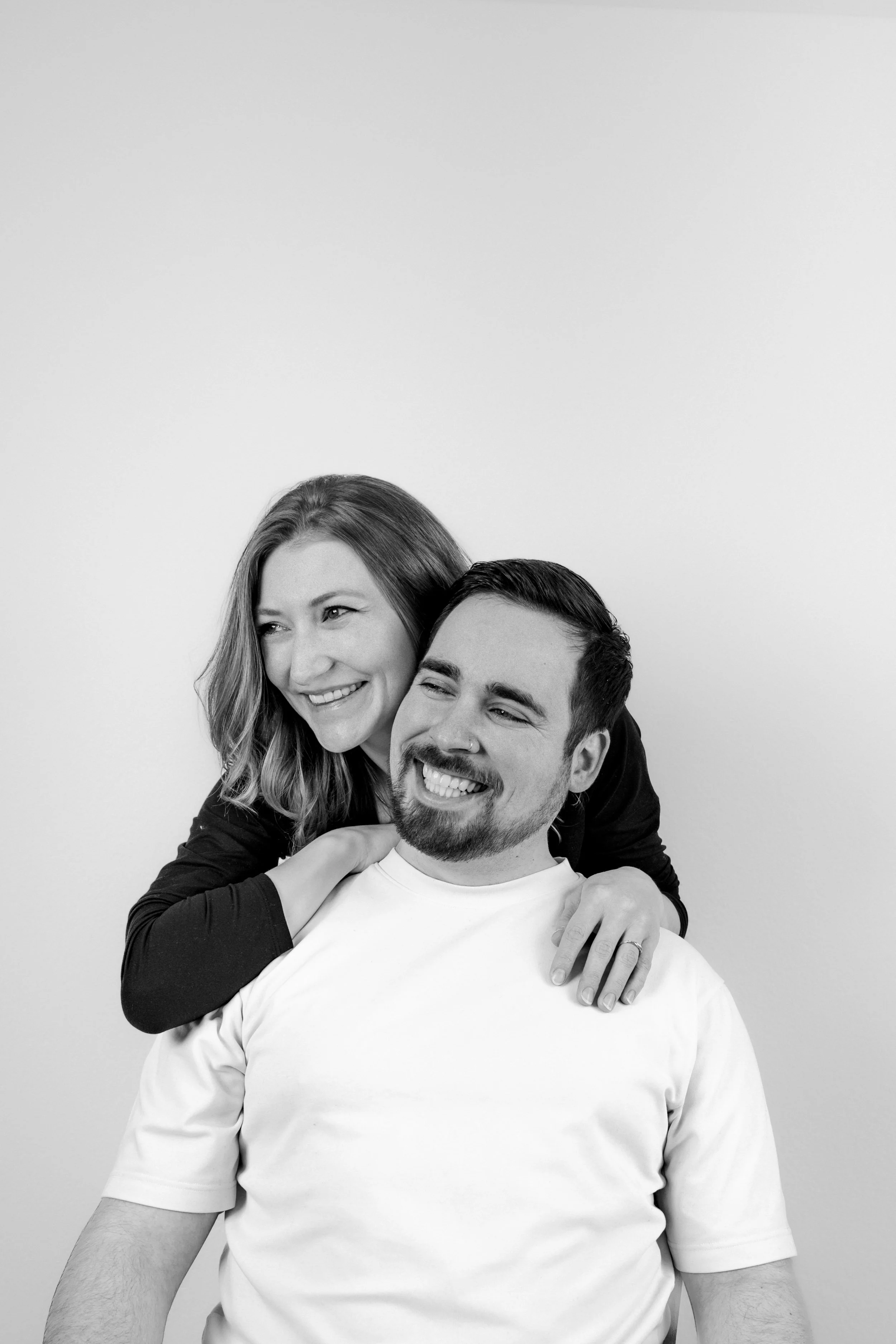 Ein lachendes Paar umarmt sich vor einfachem weißen Hintergrund, der Mann sitzt und trägt ein weißes T-Shirt, die Frau steht dahinter und legt die Arme um ihn, beide zeigen Freude und Verbundenheit.