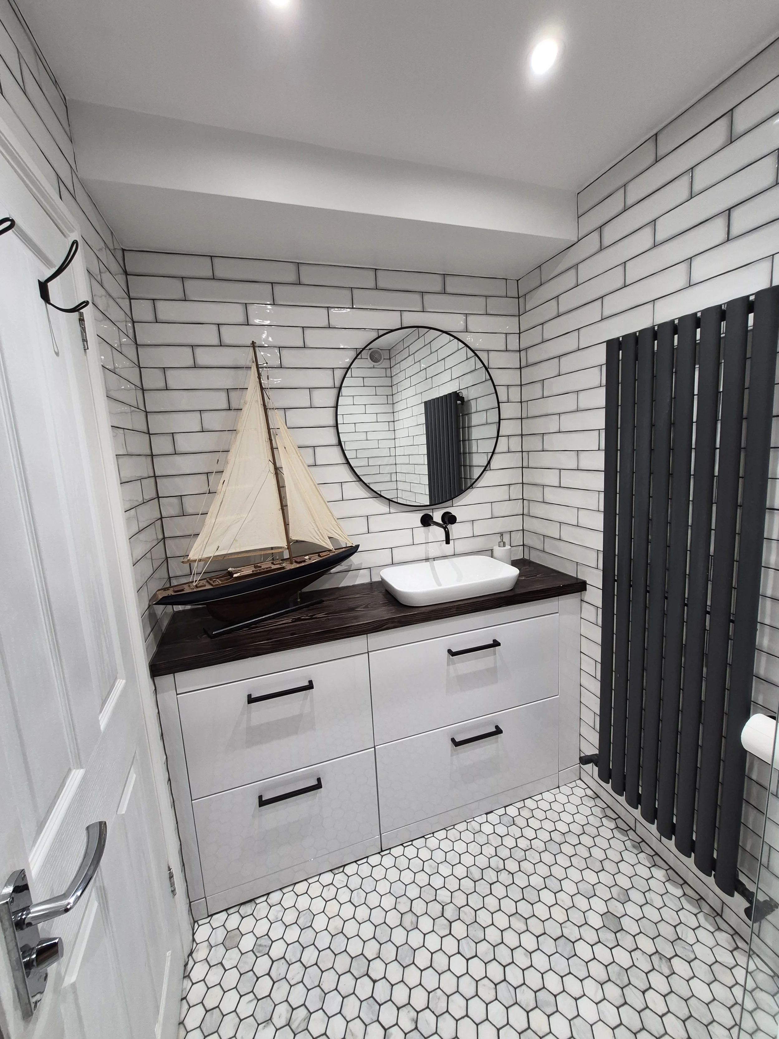 Modern bathroom with white subway tile walls, hexagon floor tiles, black radiator, and a white vanity with dark wood countertop. Decor includes a model sailboat and a round mirror.
