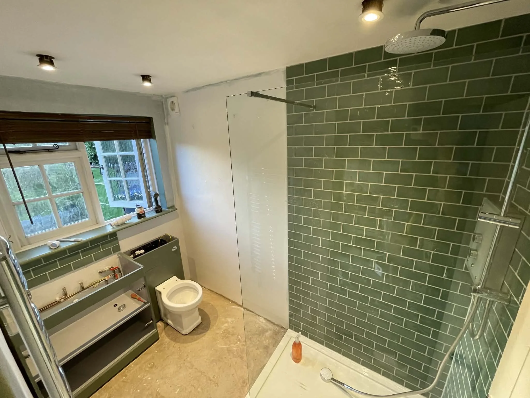 Bathroom with green tiled shower area, a window with blinds, a toilet, and a cabinet under the window, with lighting and construction tools.