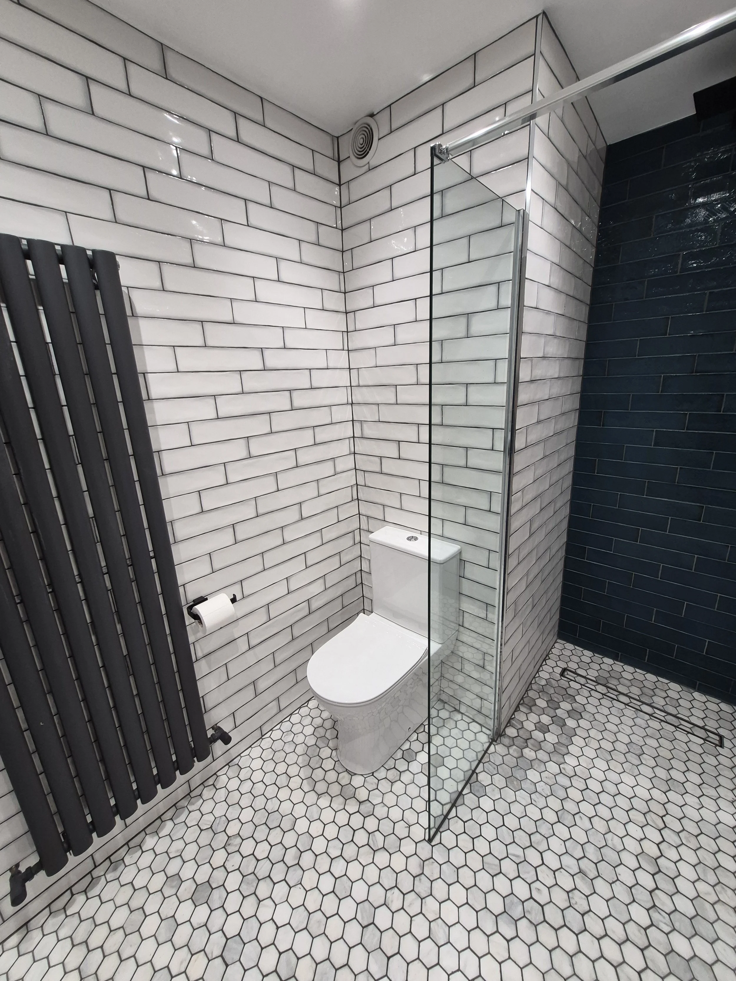 Modern bathroom with white subway tile walls, hexagon tile floor, a wall-mounted black radiator, a white toilet, and a glass shower enclosure with black grout.