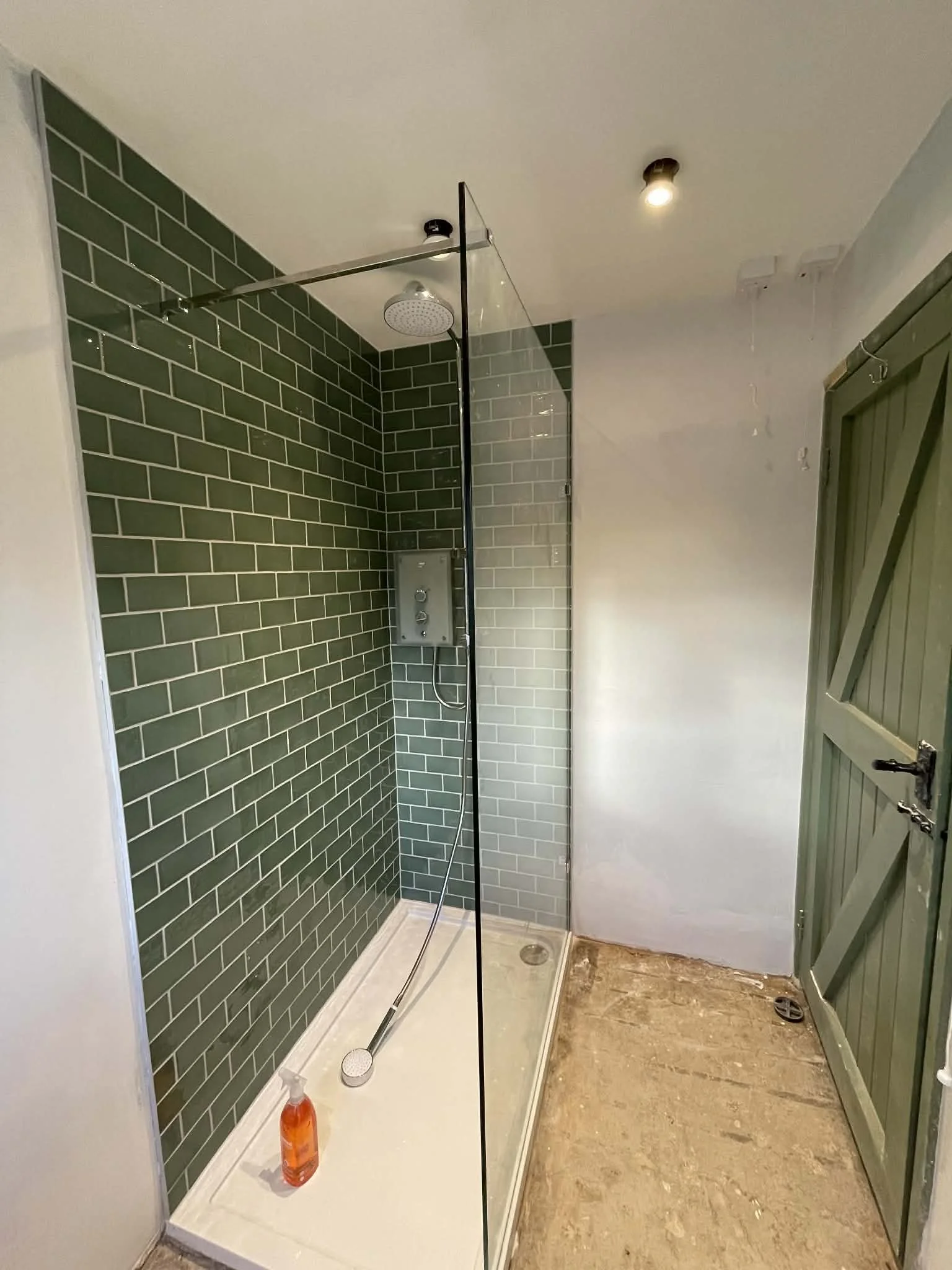 Empty shower with green subway tiles, glass partition, overhead showerhead, and a cleaning spray bottle on the shower floor. Green wooden door on the right, unfinished floor and ceiling.