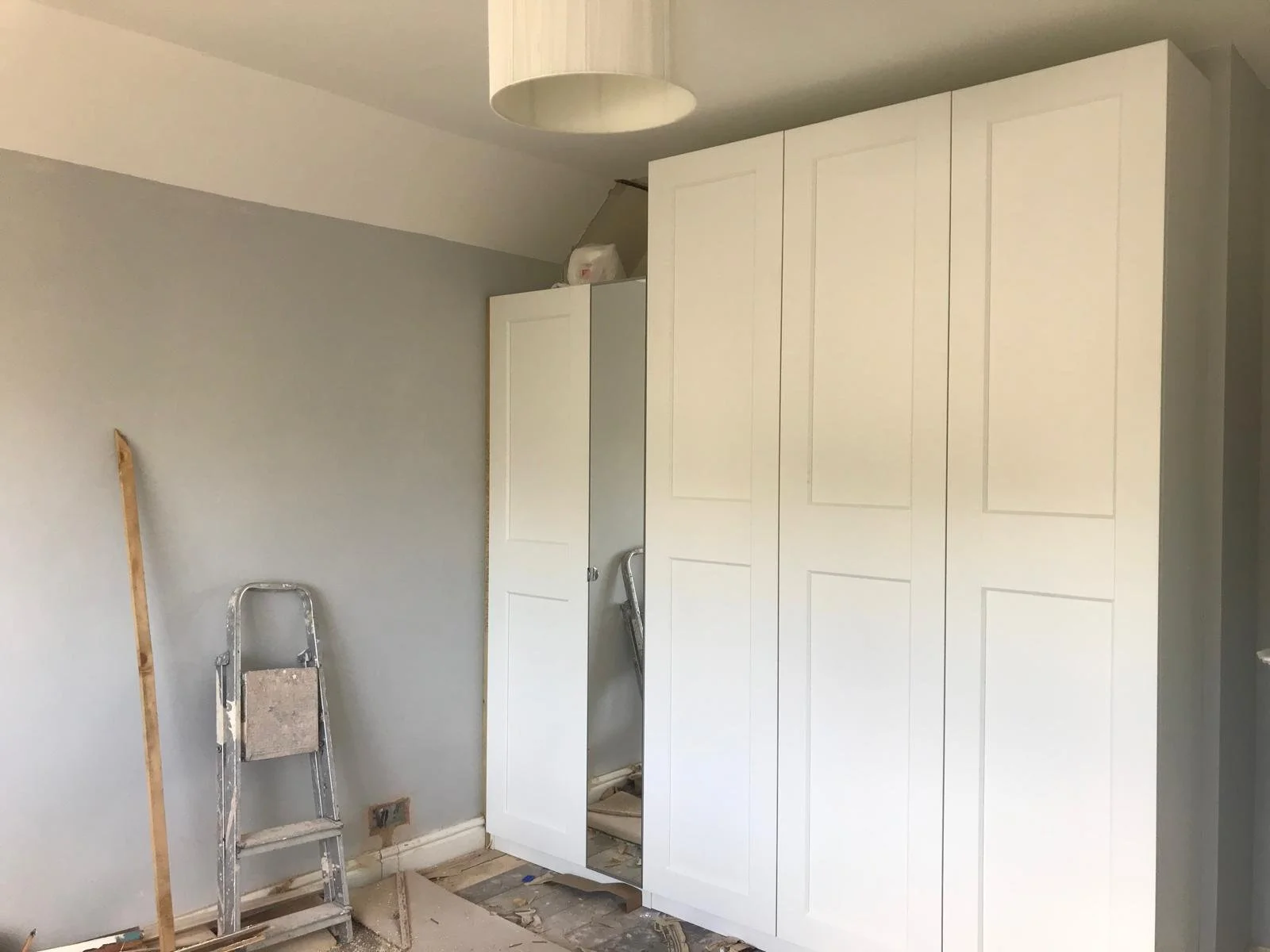 An unfinished room with a large white wardrobe, a ladder, a wooden stick, and construction debris on the floor.