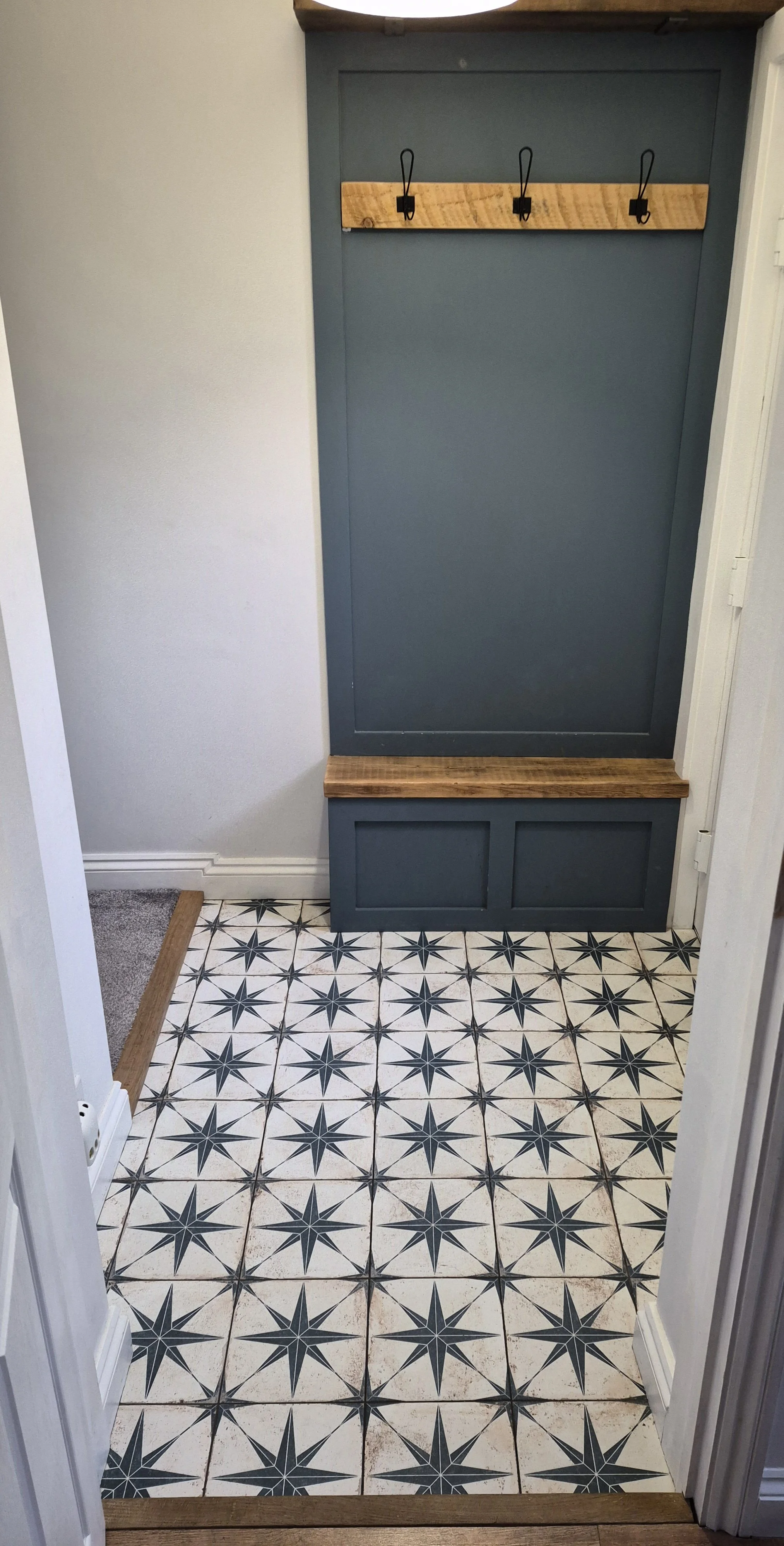 A hallway with white walls, a patterned floor with star and diamond motifs, and a blue panel with a wooden bench and three hook hangers.