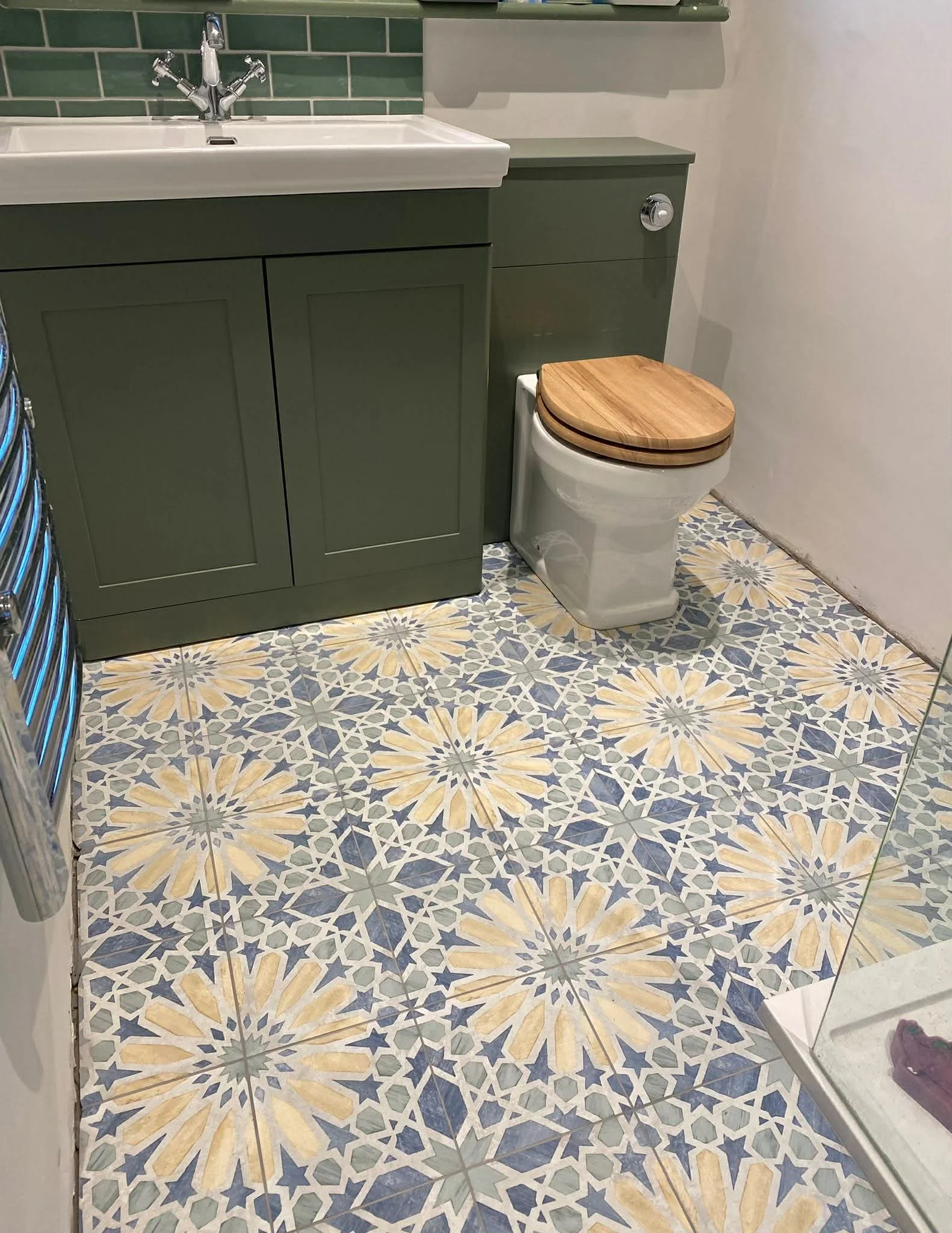Close-up of a bathroom with patterned tile floor, green vanity with sink, and a toilet with a wooden seat cover.