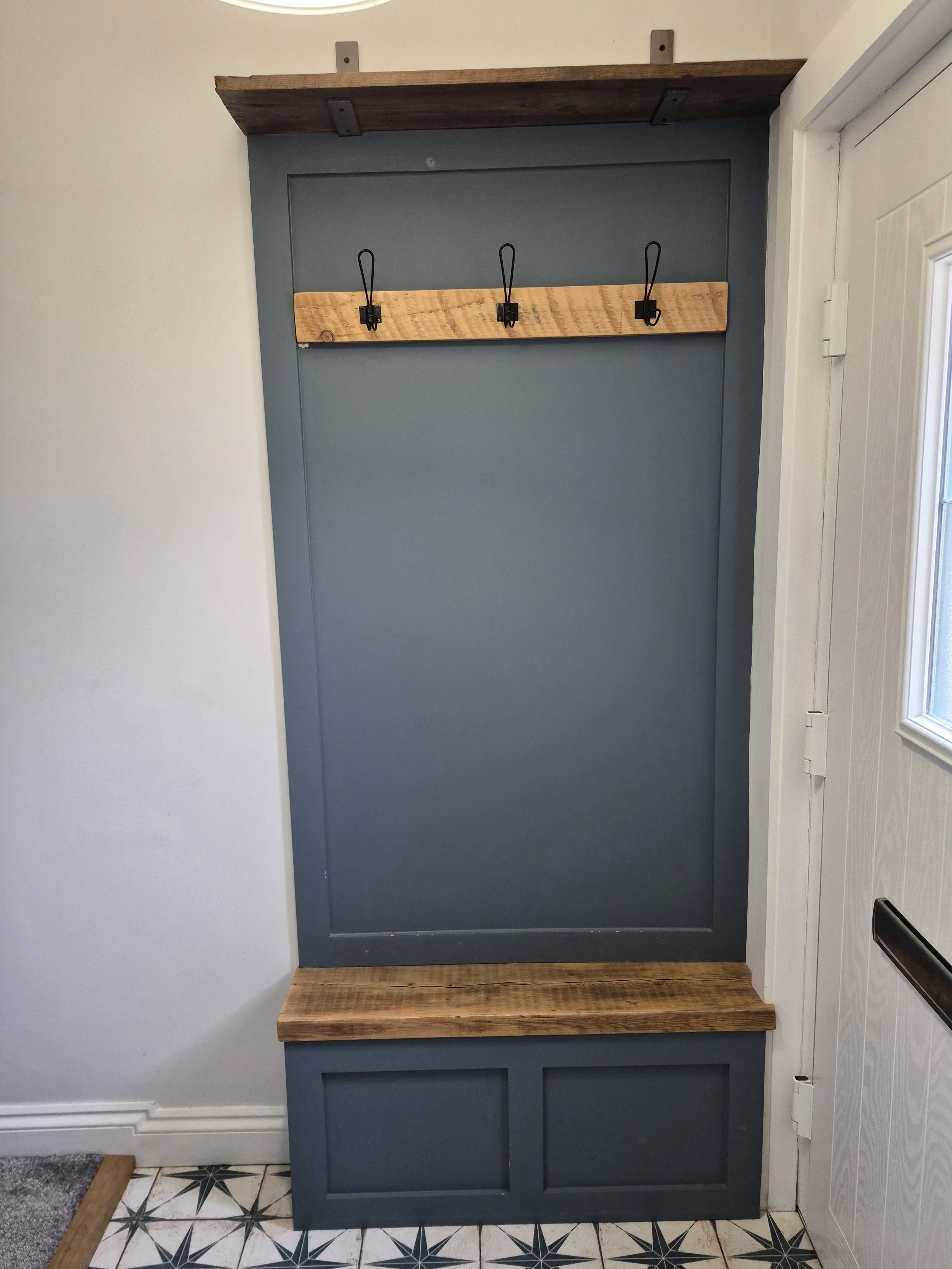A wooden coat rack with three hooks mounted on a blue wall near a white door, with a wooden bench below and an overhanging shelf above.