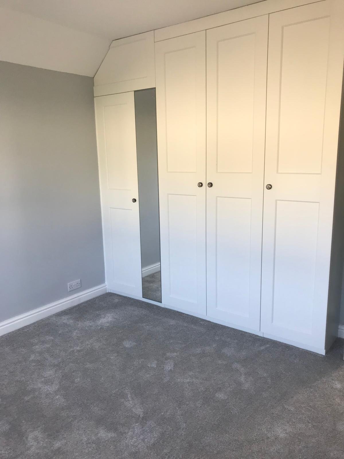 Empty bedroom corner with built-in closet featuring white sliding doors, one with a full-length mirror, and a grey wall with electrical outlet and carpeted floor.