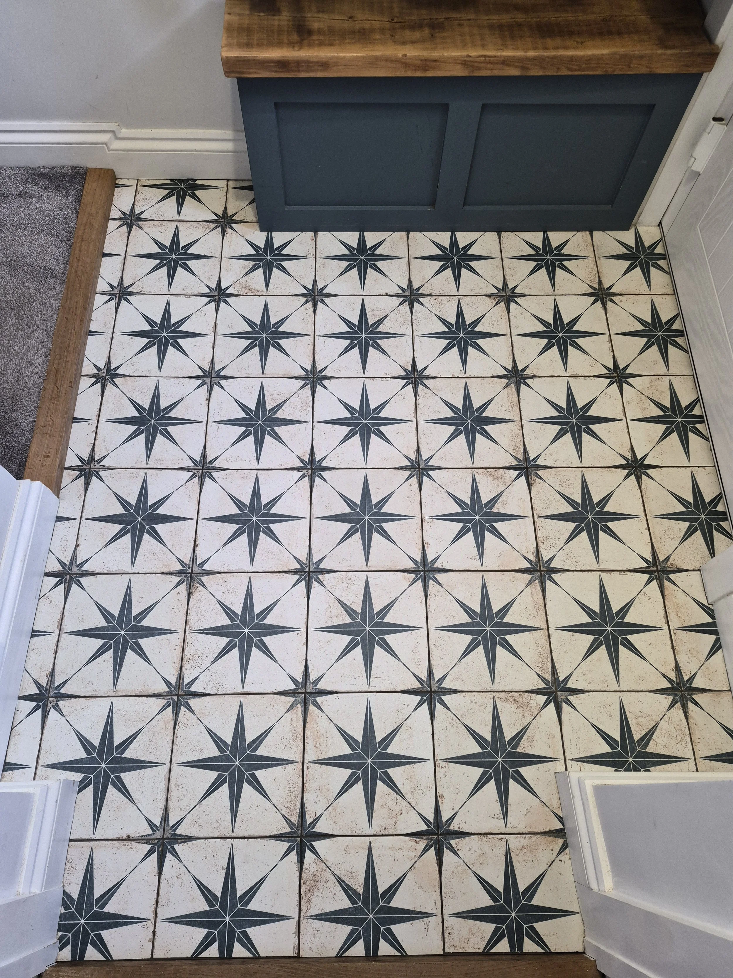 Patterned floor tiles with black starburst designs on a white background in a room corner, with a blue cabinet and wood trim.