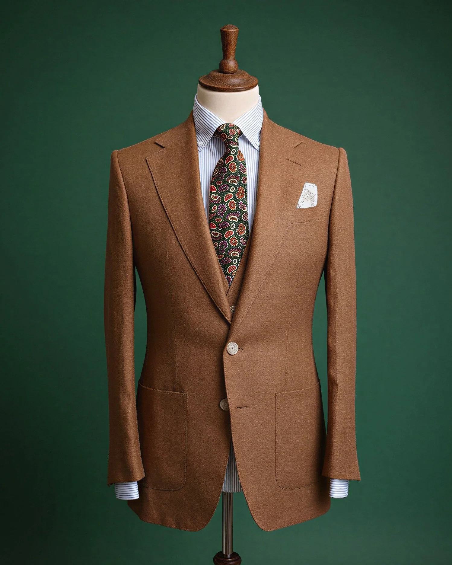 A brown suit jacket on a mannequin with a white and blue striped dress shirt, a colorful paisley tie, and a white pocket square against a green background.