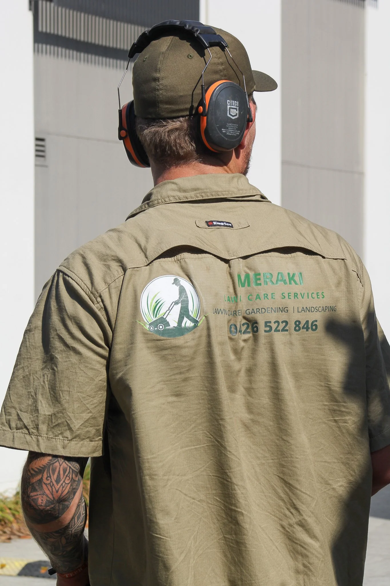 Back view of a man wearing a khaki work shirt and a matching cap, with soundproof earmuffs. The shirt has a logo and contact number for Meraki Lawn Care Services, which offers lawn care, gardening, and landscaping. The man has a tattoo on his left arm and is outdoors in front of a building