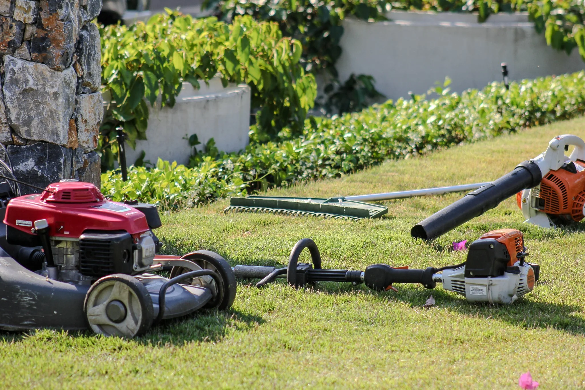 Lawn equipment including a leaf blower, trimmer, and weed cutter on a well-maintained garden lawn with green bushes and a stone wall in the background. - Meraki Lawn Care Services