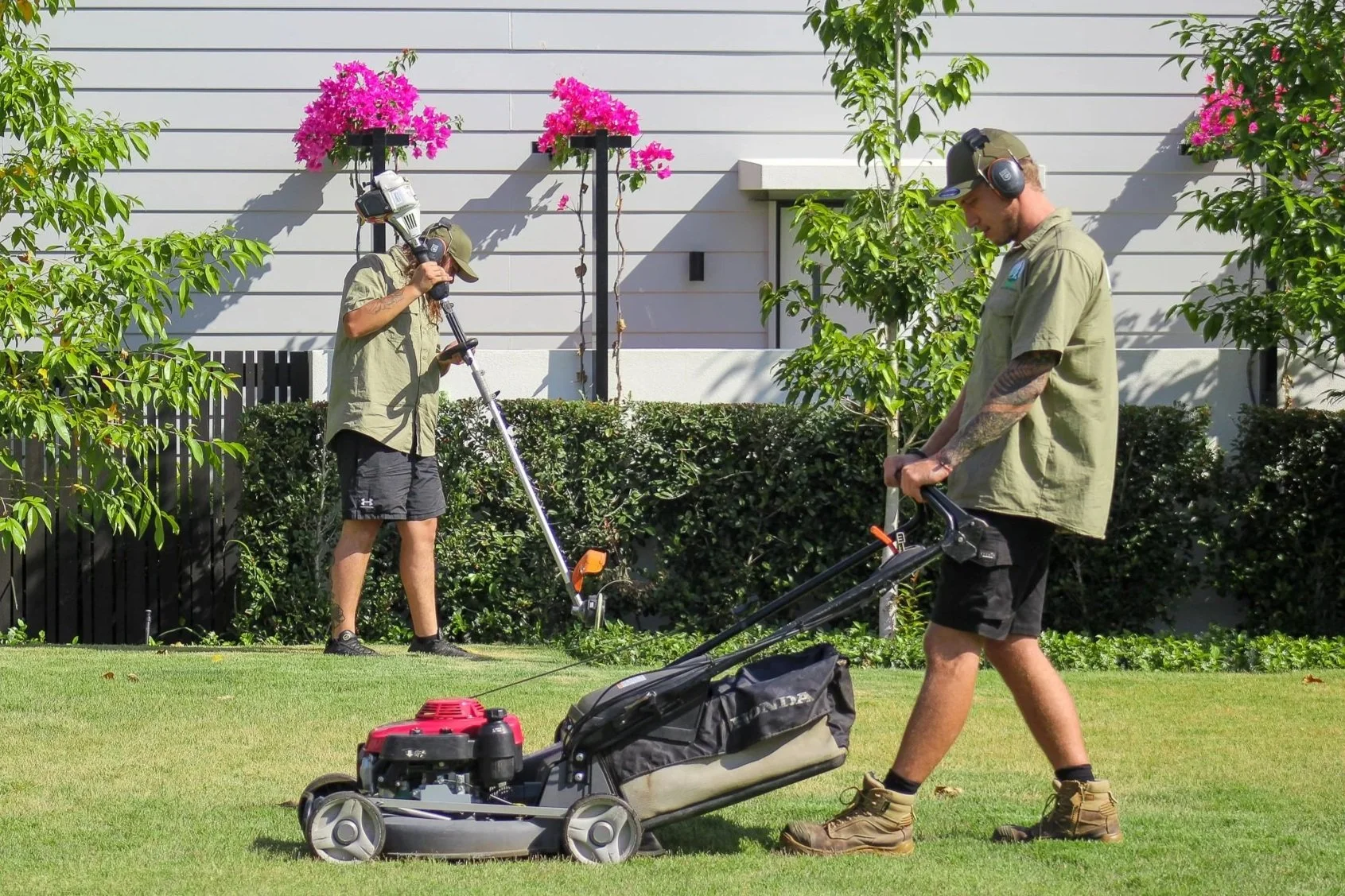 Two men wearing khaki shirts, black shorts, and brown work boots in a garden; one is pushing a lawnmower while the other is using a ground-penetrating metal detector. - Meraki Lawn Care Services