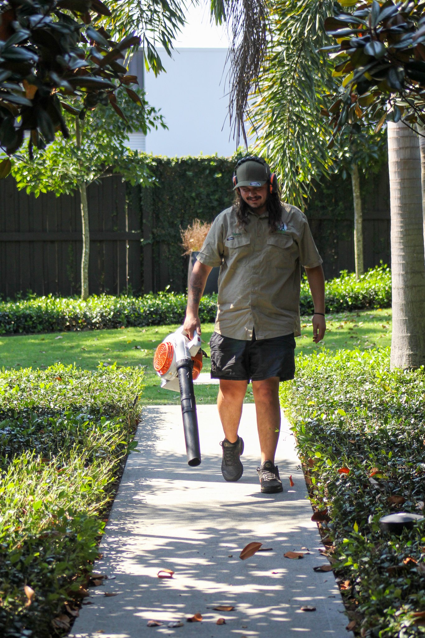Man wearing headphones and sunglasses, operating leaf blower on a sidewalk in a garden with lush bushes and trees. - Meraki Lawn Care Services
