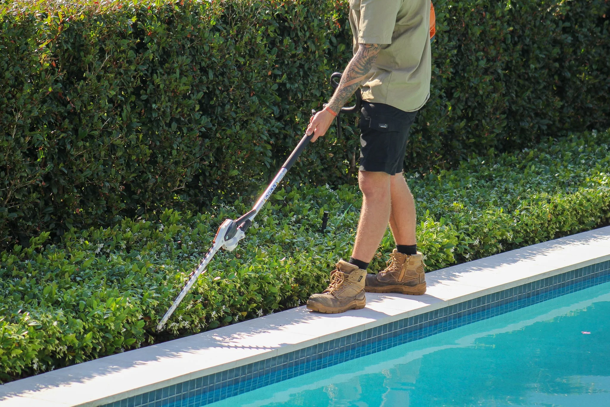 Person trimming bushes along a swimming pool using a hedge trimmer. - Meraki Lawn Care Services