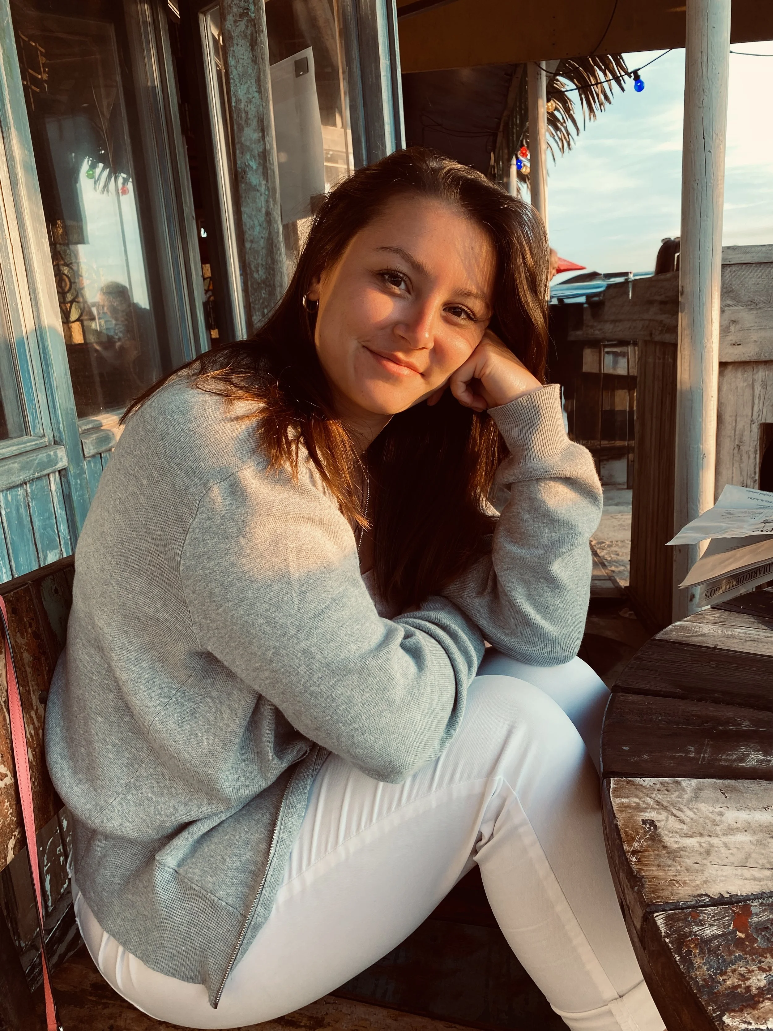 Junge Frau mit dunklem Haar sitzt draußen an einem runden Holz-Tisch, trägt einen grauen Pullover und weiße Hosen, lächelt bei Sonnenuntergang, im Hintergrund Holzwände und bunte Lichter.