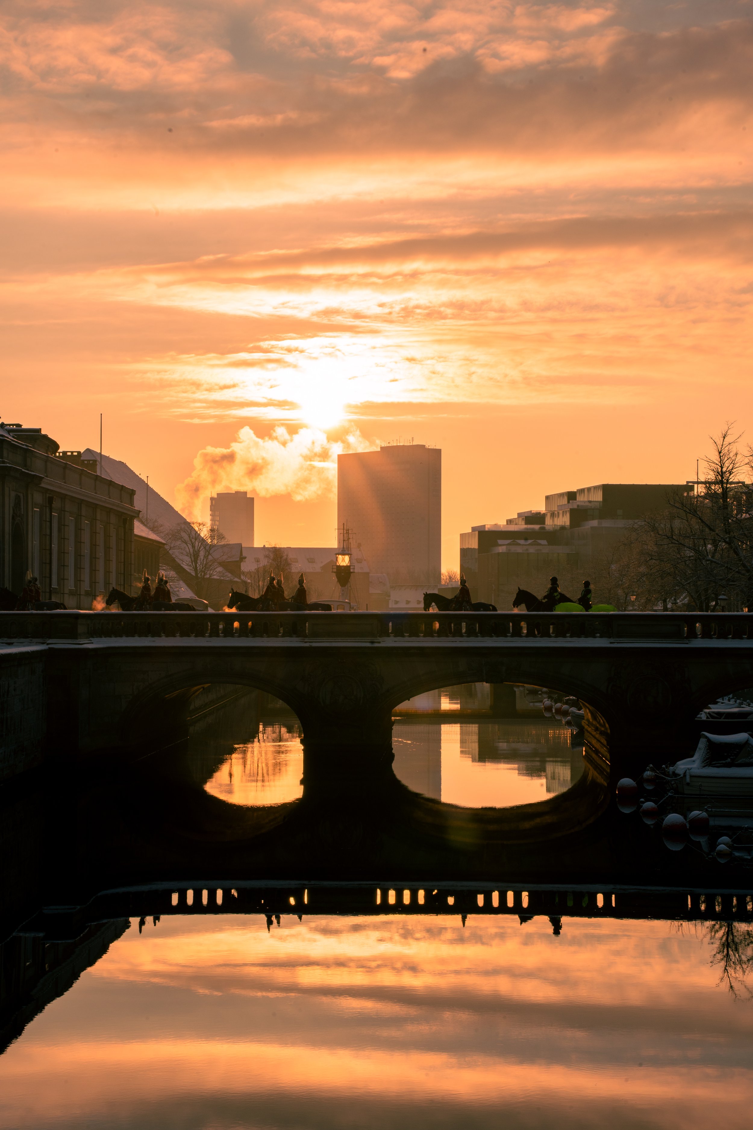 Sunset over a city river, with a bridge and reflections in the water, with silhouetted buildings and trees.