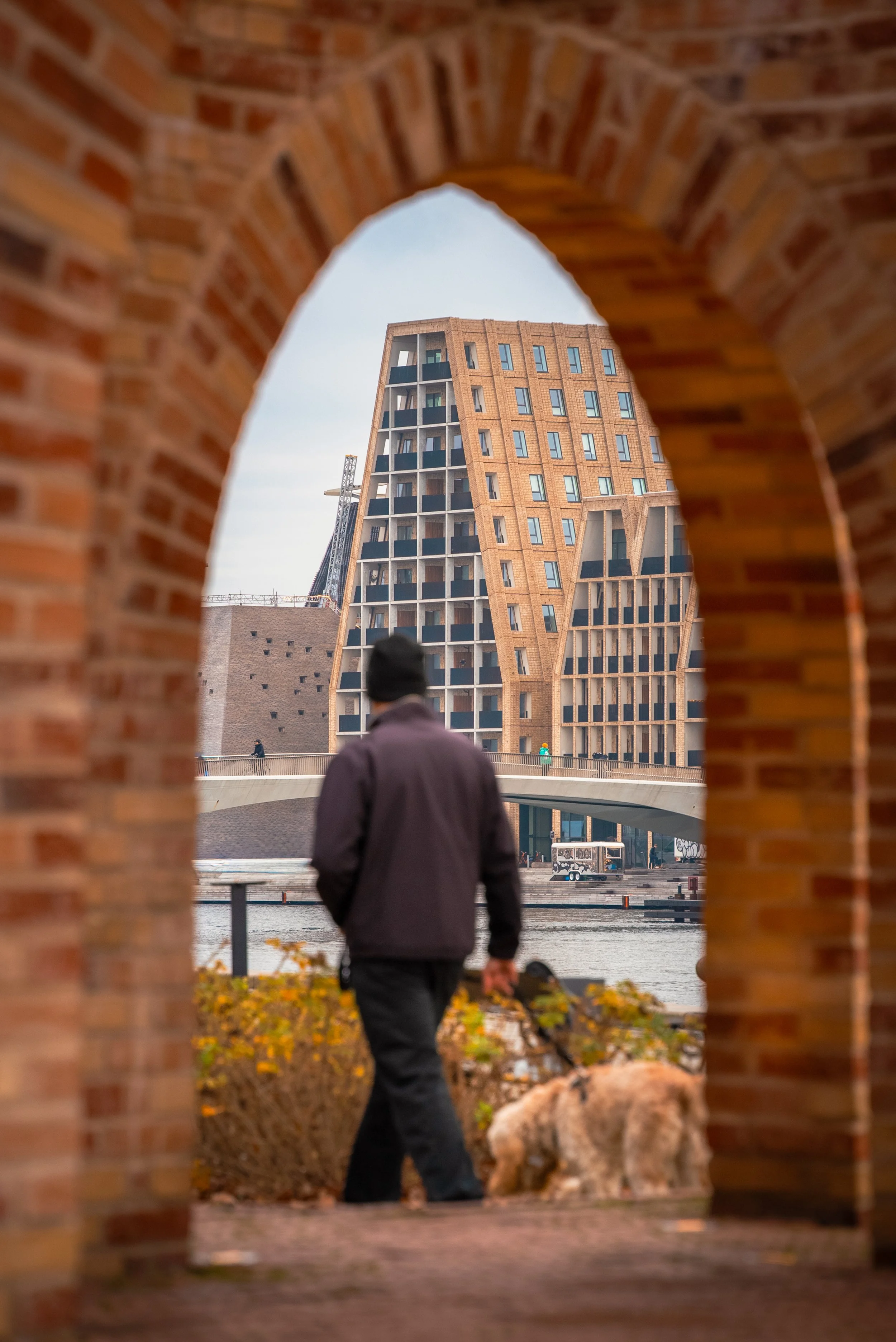 Man walking a dog near a river, viewed through a brick archway with a modern building in the background.