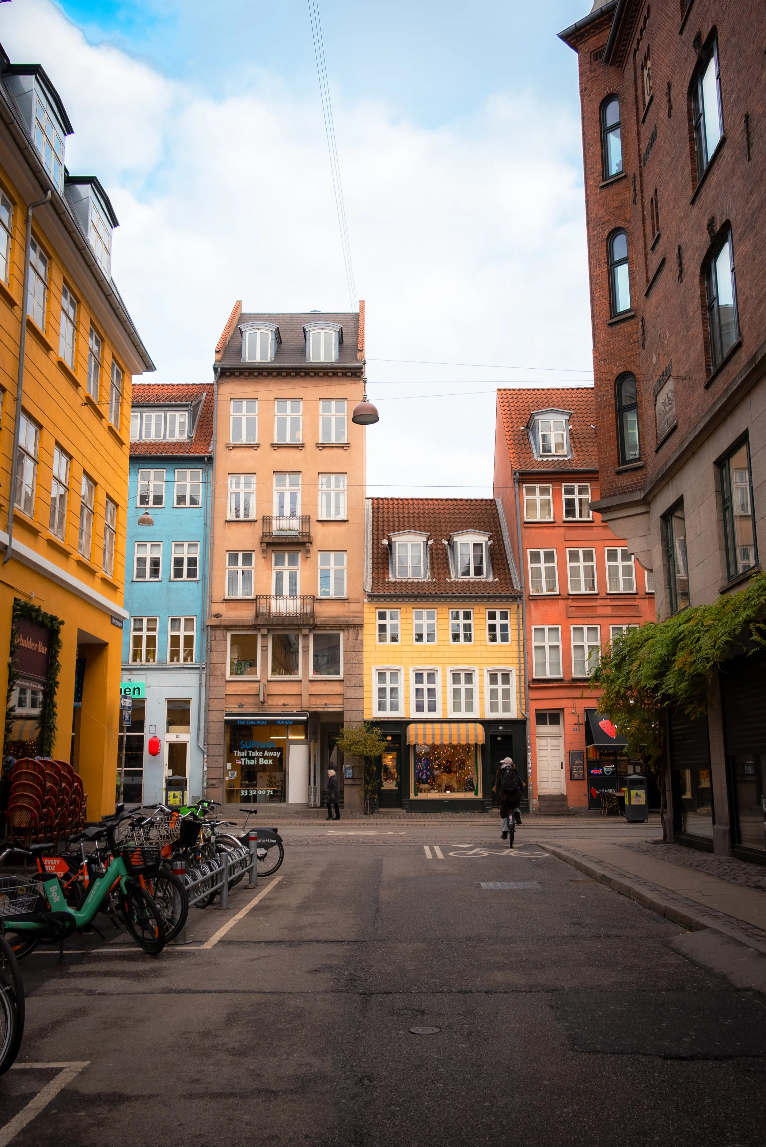 City street with colorful apartment buildings, parked bicycles, pedestrians, and a shop with a striped awning.