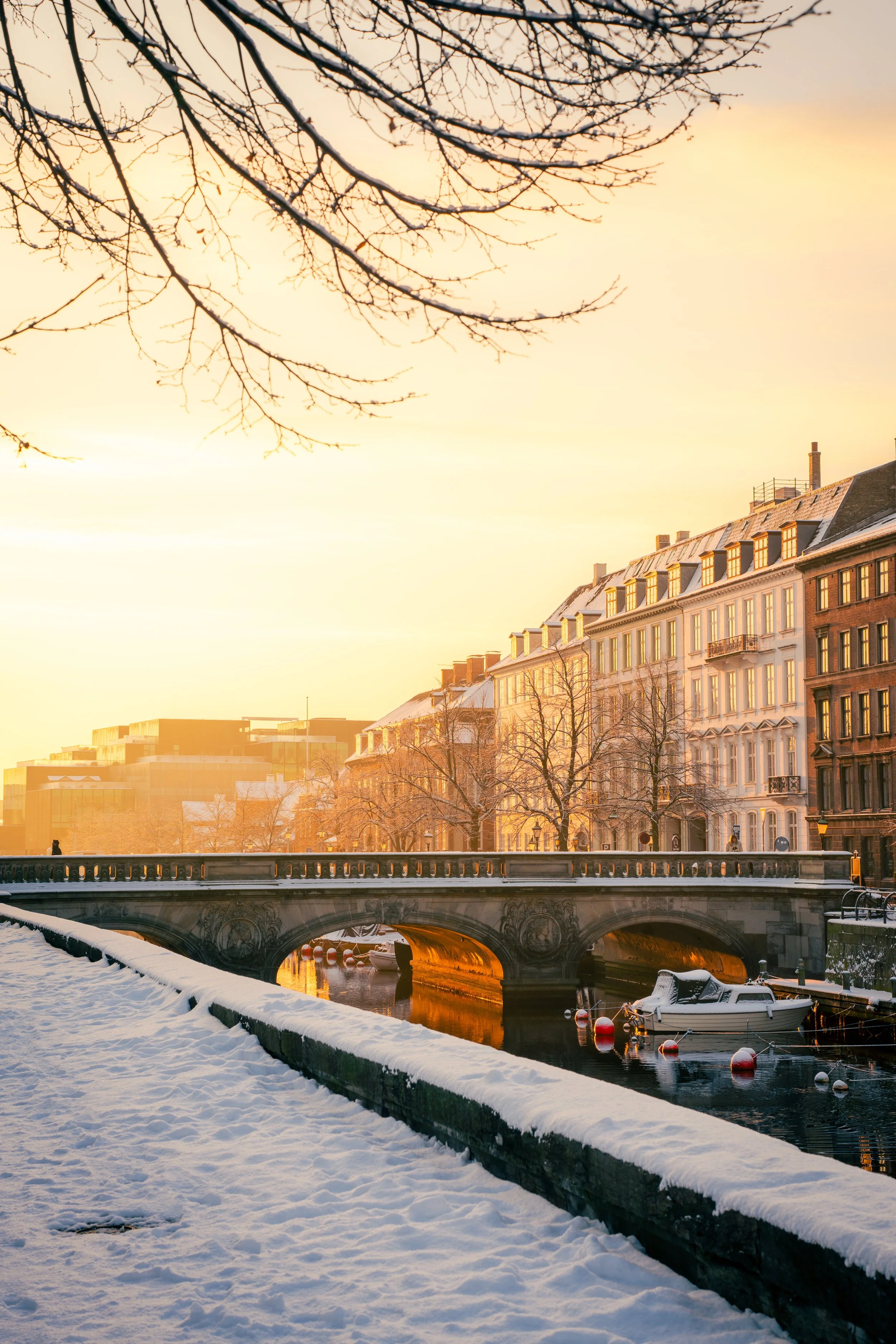 A snow-covered riverbank with a bridge reflected on the water, rowboats docked, and buildings with snow on their roofs under a sunset sky and bare tree branches.