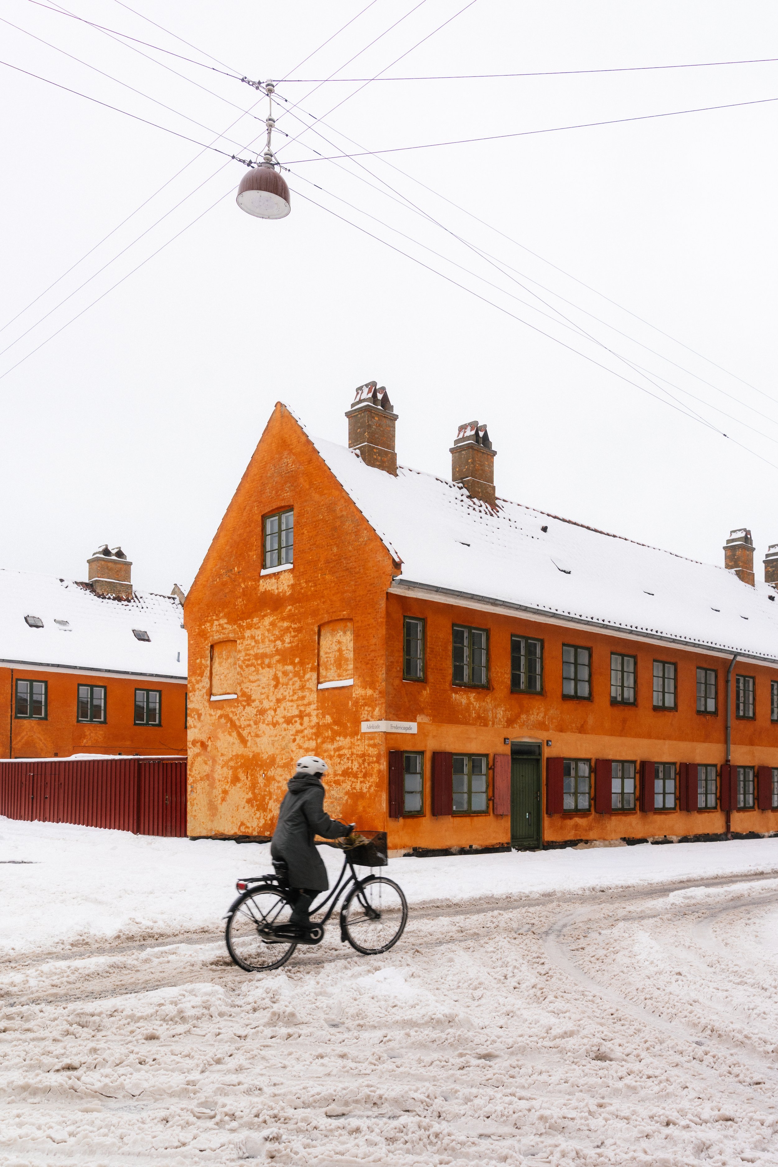 A person wearing a helmet rides a bicycle on a snow-covered street in front of an orange building with multiple windows and chimneys, under overhead electric wires on a cloudy winter day.