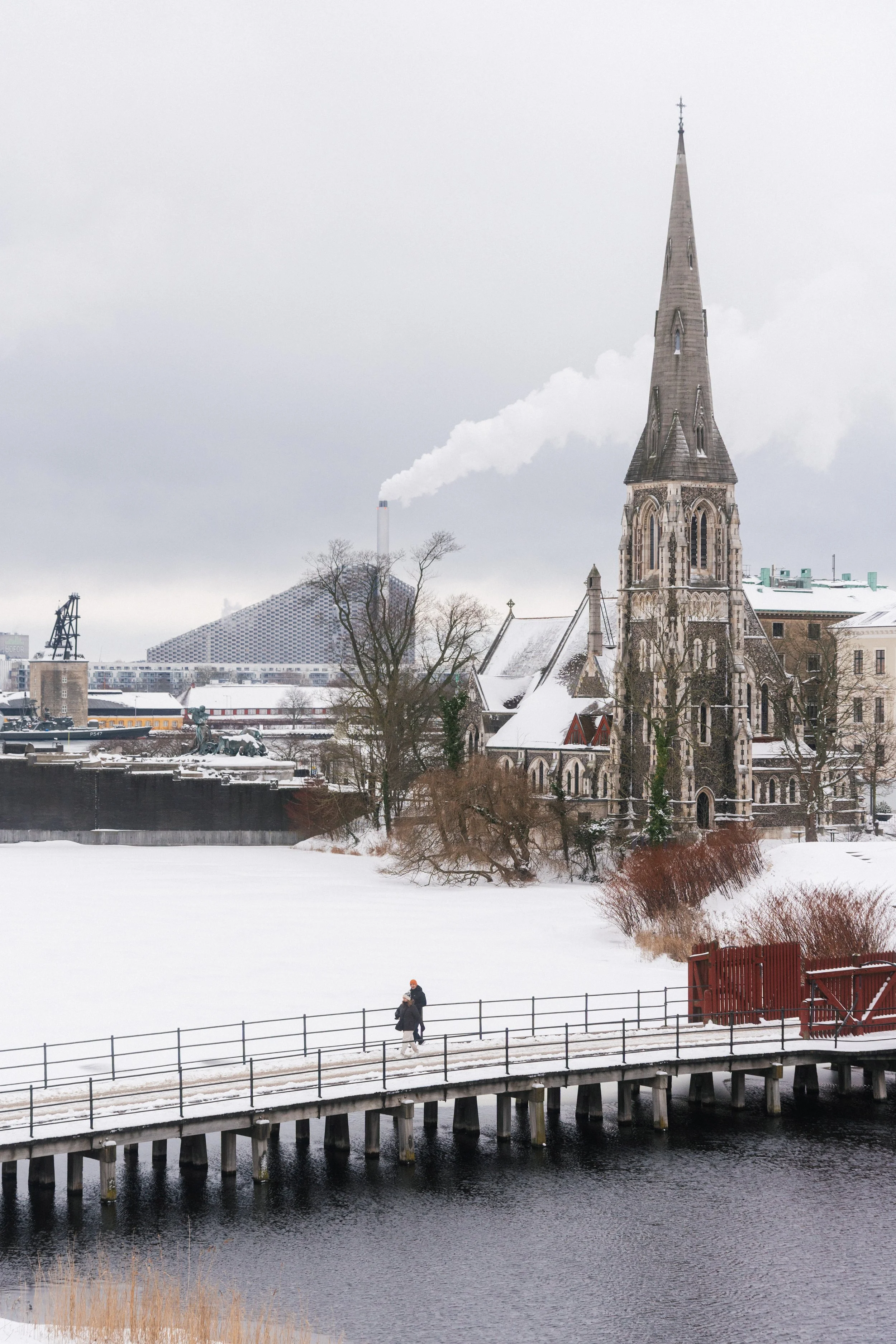 A snowy scene of a city with a church in the foreground, a modern building in the background, and a bridge over a river with two people walking on it.