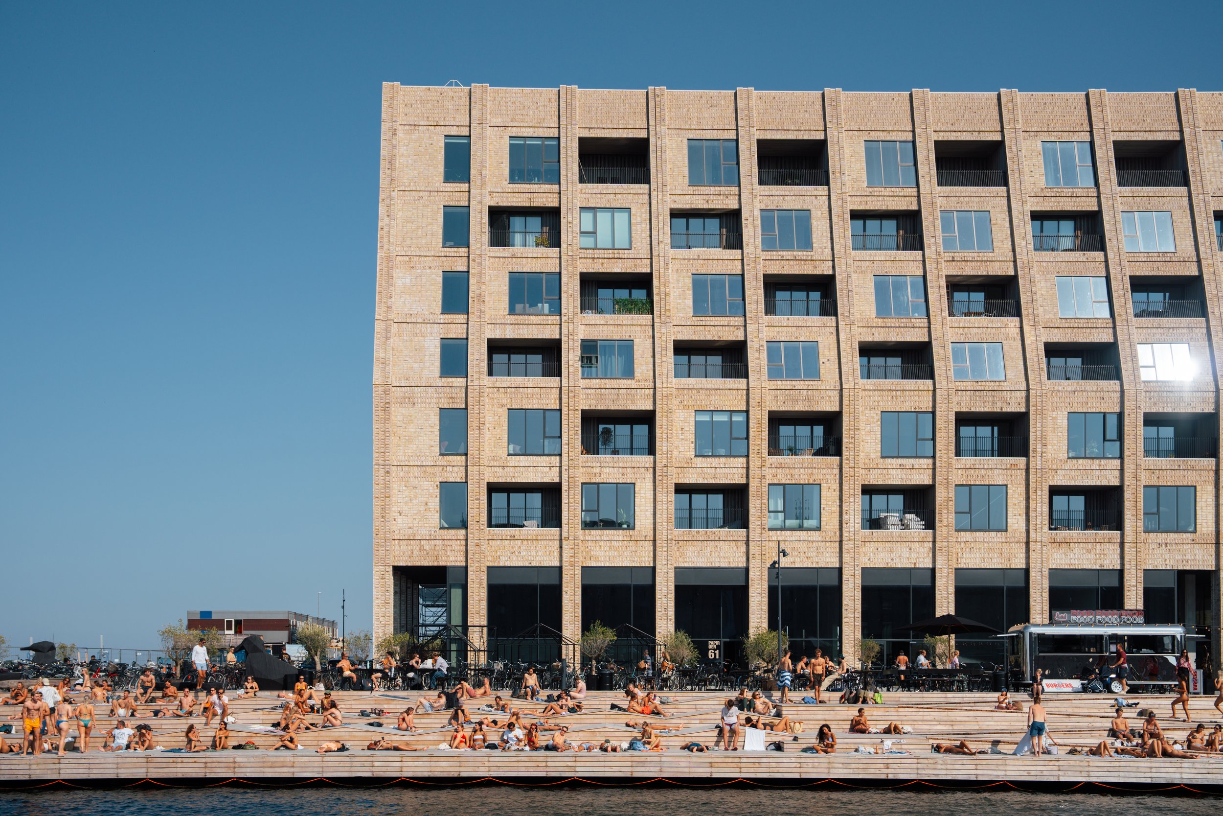 People swimming and lounging on the steps at a waterfront pool in front of a large beige brick apartment building with many windows and balconies under a clear blue sky.