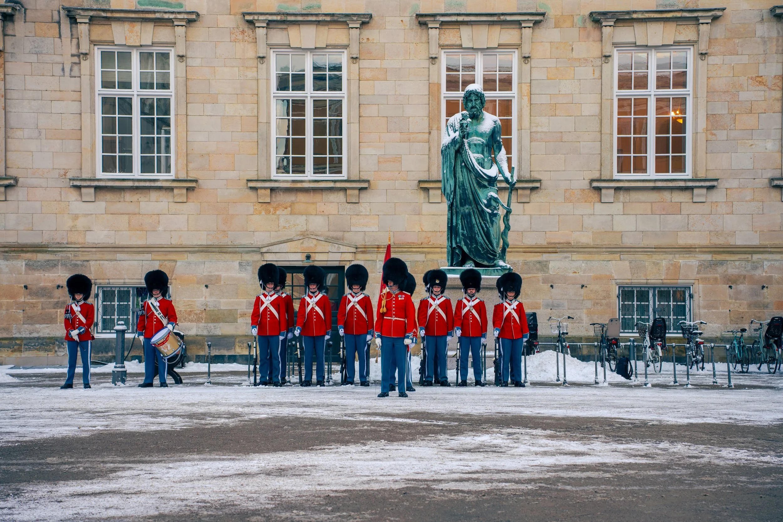 A group of Royal Canadian Mounted Police officers dressed in red uniforms and bearskin hats standing in formation with a drummer in front, in front of a stone building with four large windows and a statue of a bearded man holding a staff in snow.