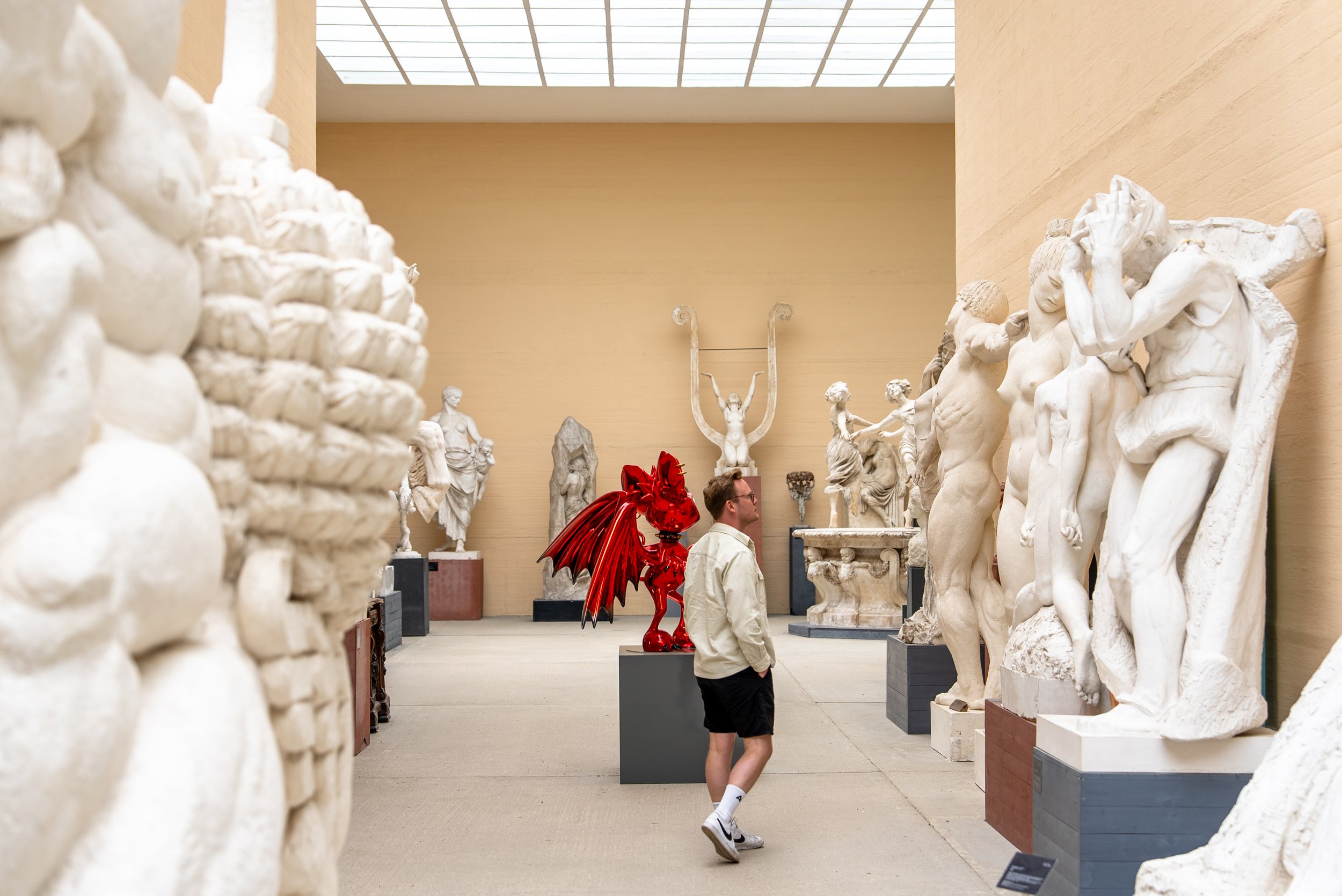 A person in a museum surrounded by marble sculptures of various figures, with a bright ceiling window overhead.